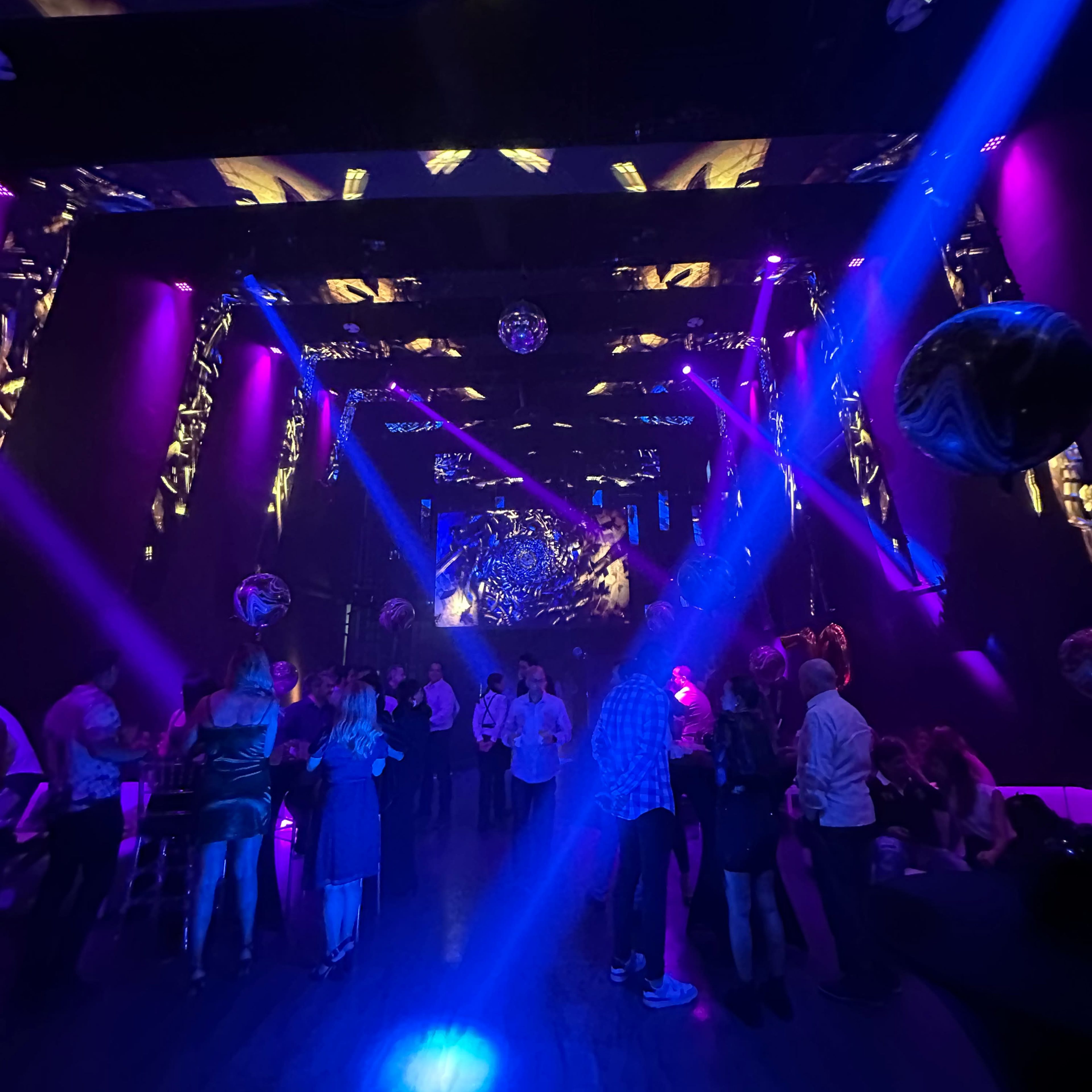 A lively nightclub scene features patrons mingling under vibrant purple and blue lighting, with beams of light cutting through the atmosphere and mirrors reflecting across the space.