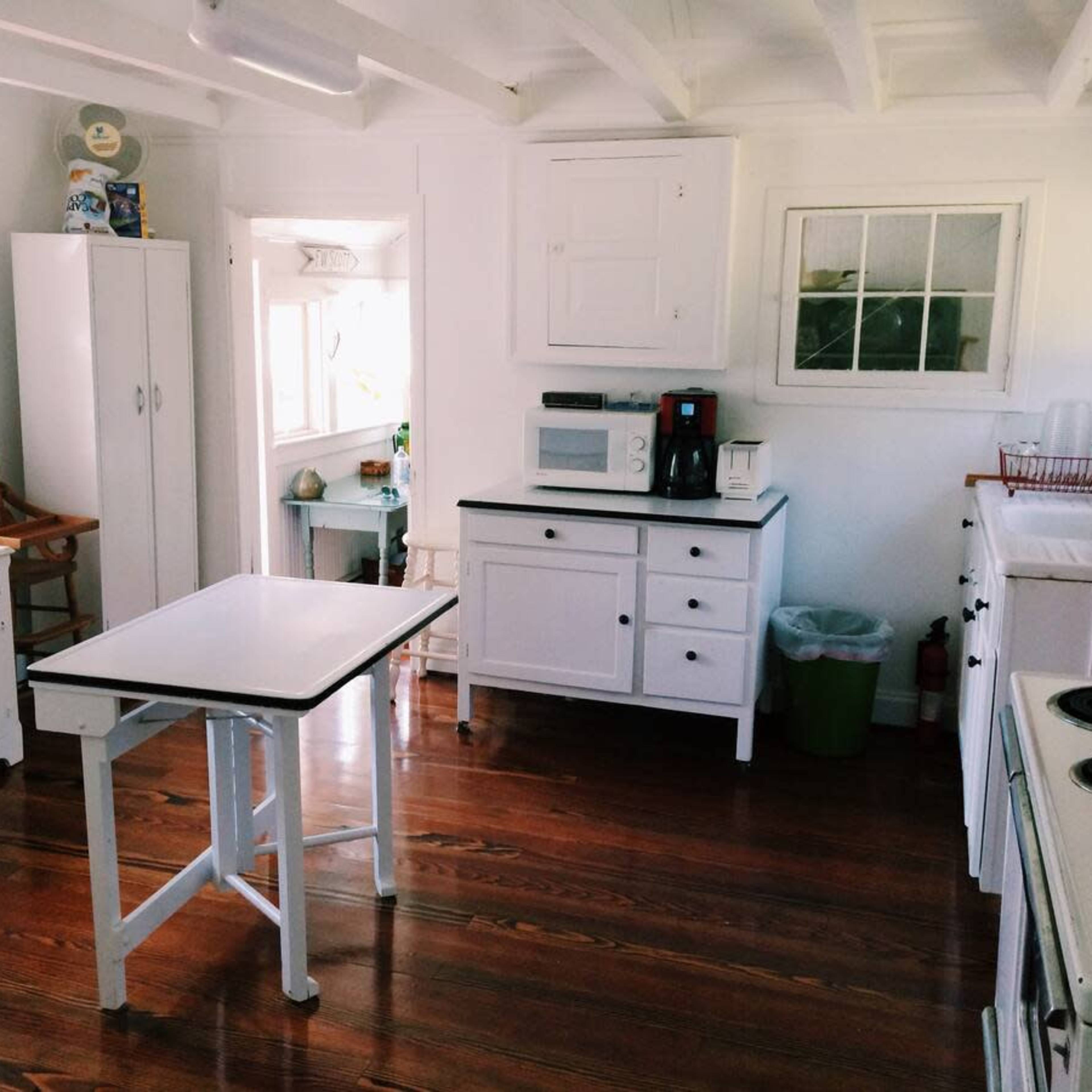 A bright kitchen with white cabinets, a table, and a coffee maker, featuring wooden floors and a window that lets in natural light.