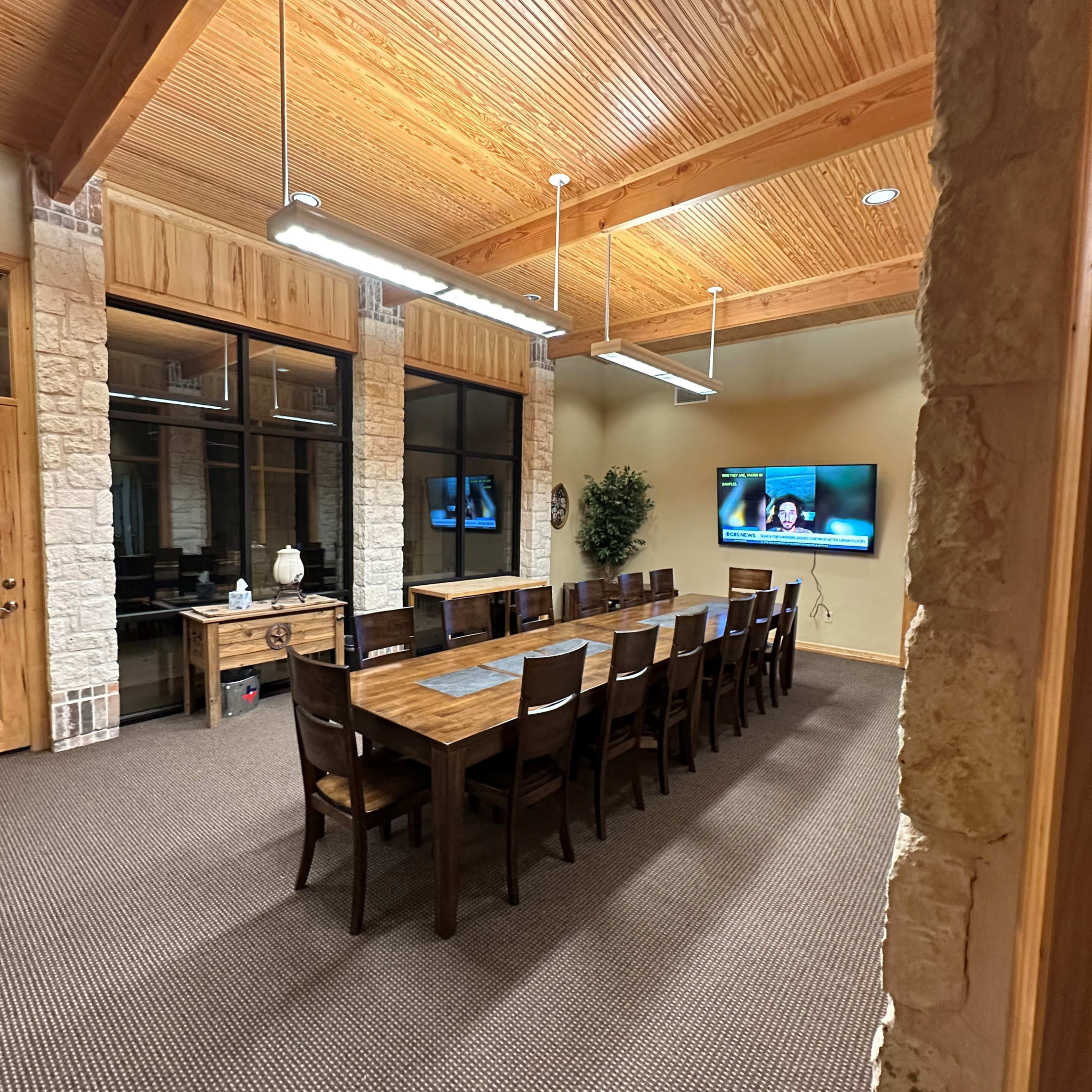A large conference room with a long wooden table, surrounded by chairs, and a wall-mounted television displaying a video call.