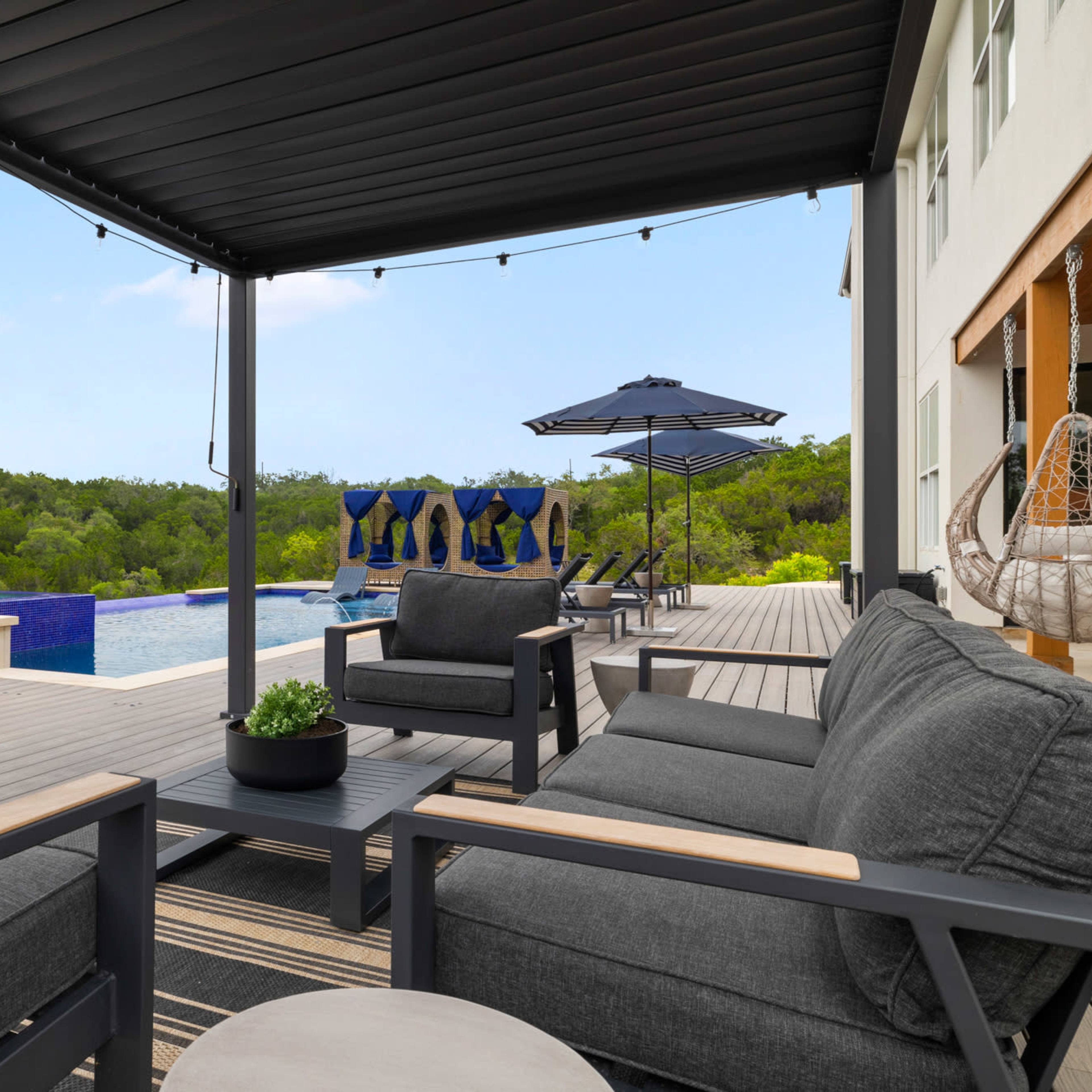 A covered patio area features dark furniture and a table, overlooking a swimming pool and lush greenery in the background.