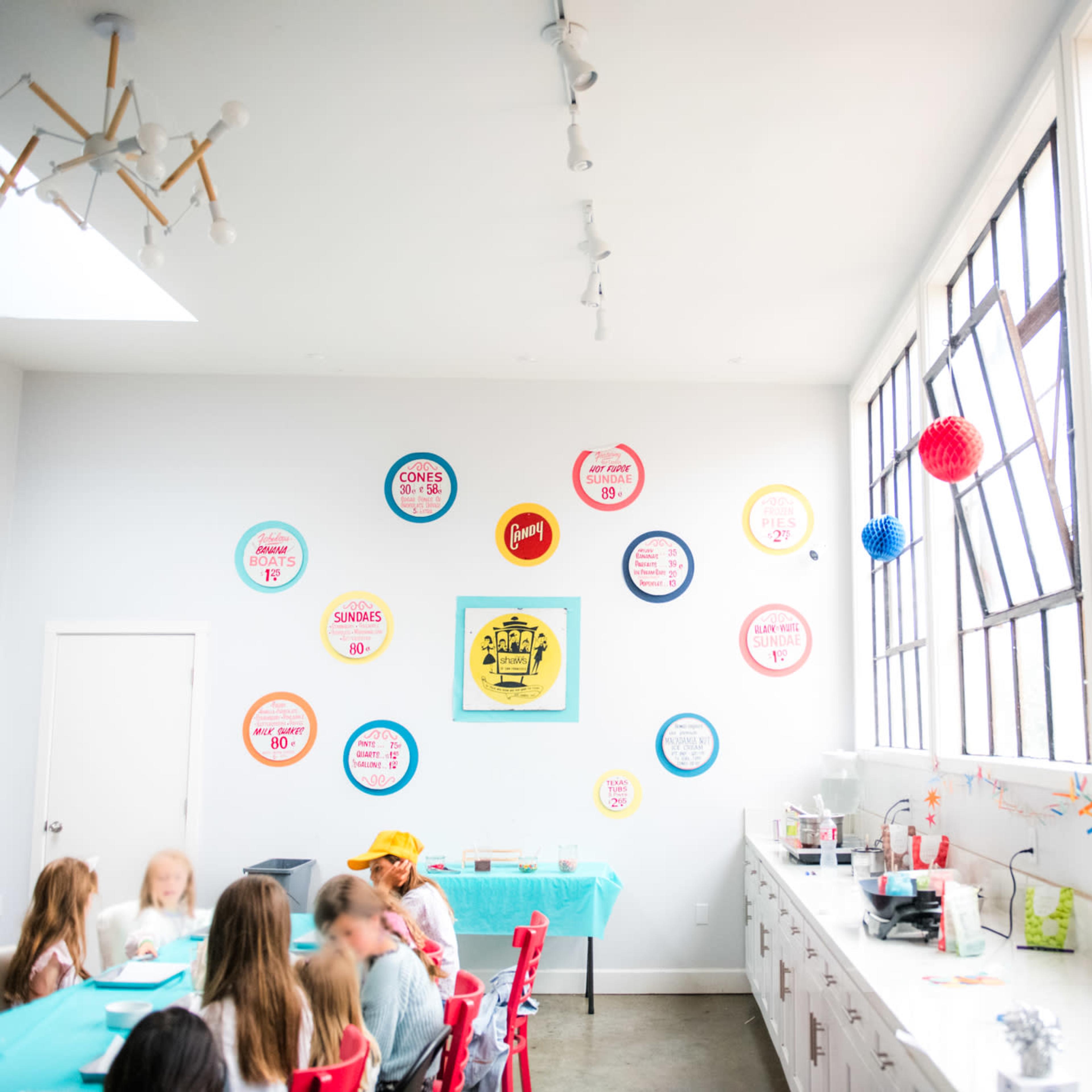 A bright, modern kitchen features a long table with children gathered around it, colorful wall decorations, and large windows allowing natural light to fill the space.