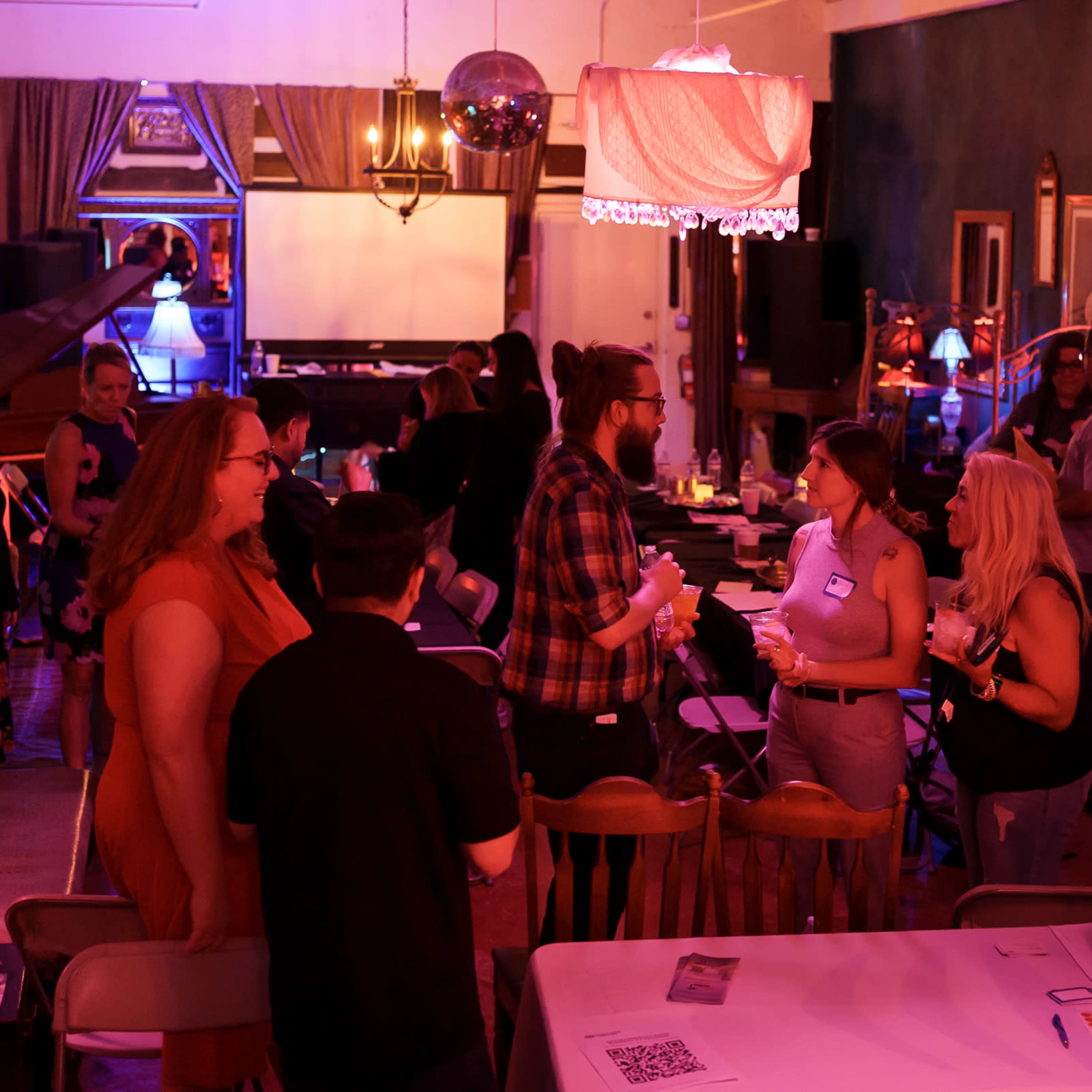 A group of people engages in conversation at a dimly lit venue with tables and decorative lighting.