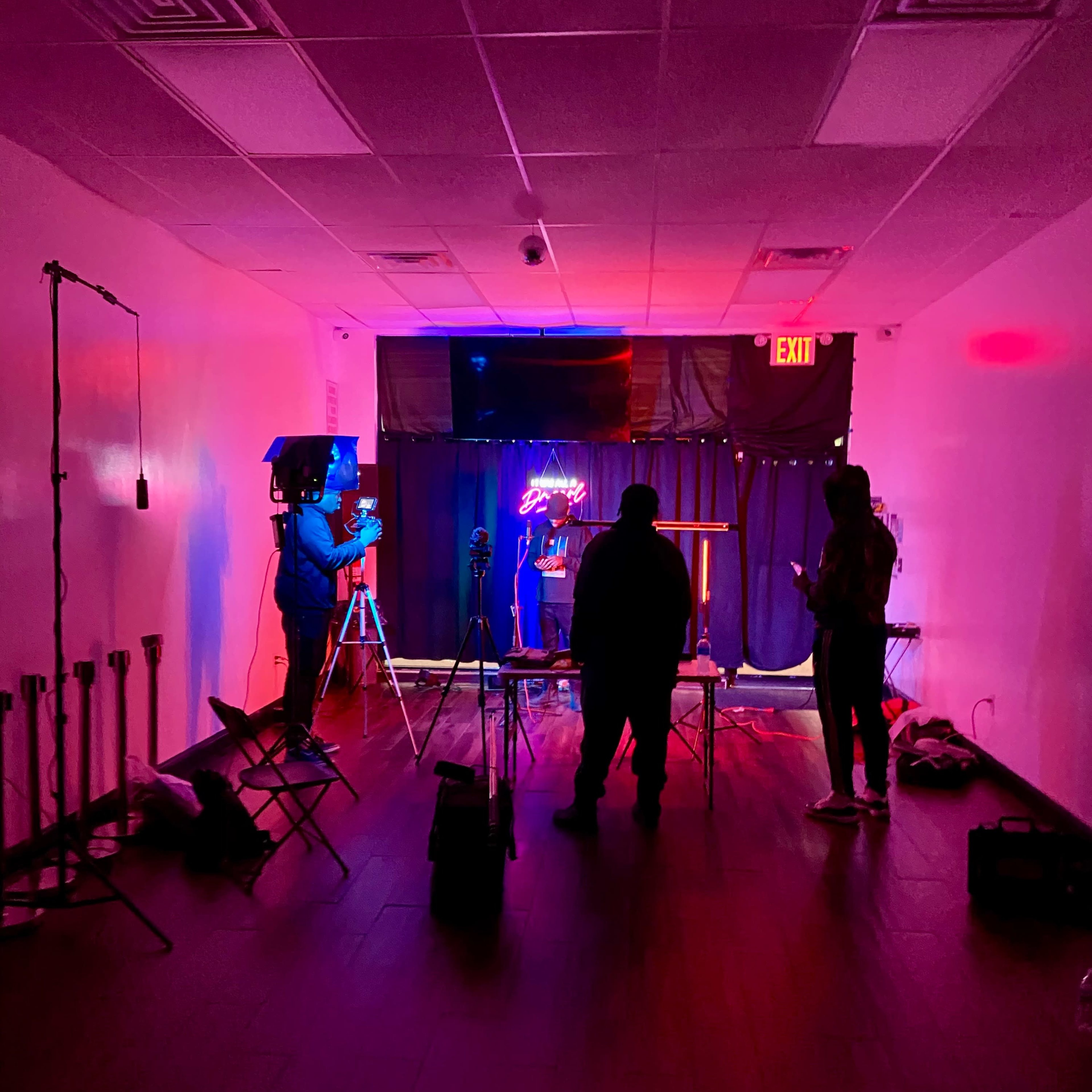 A dimly lit room with three people silhouetted against colorful lighting, preparing equipment for a shoot.