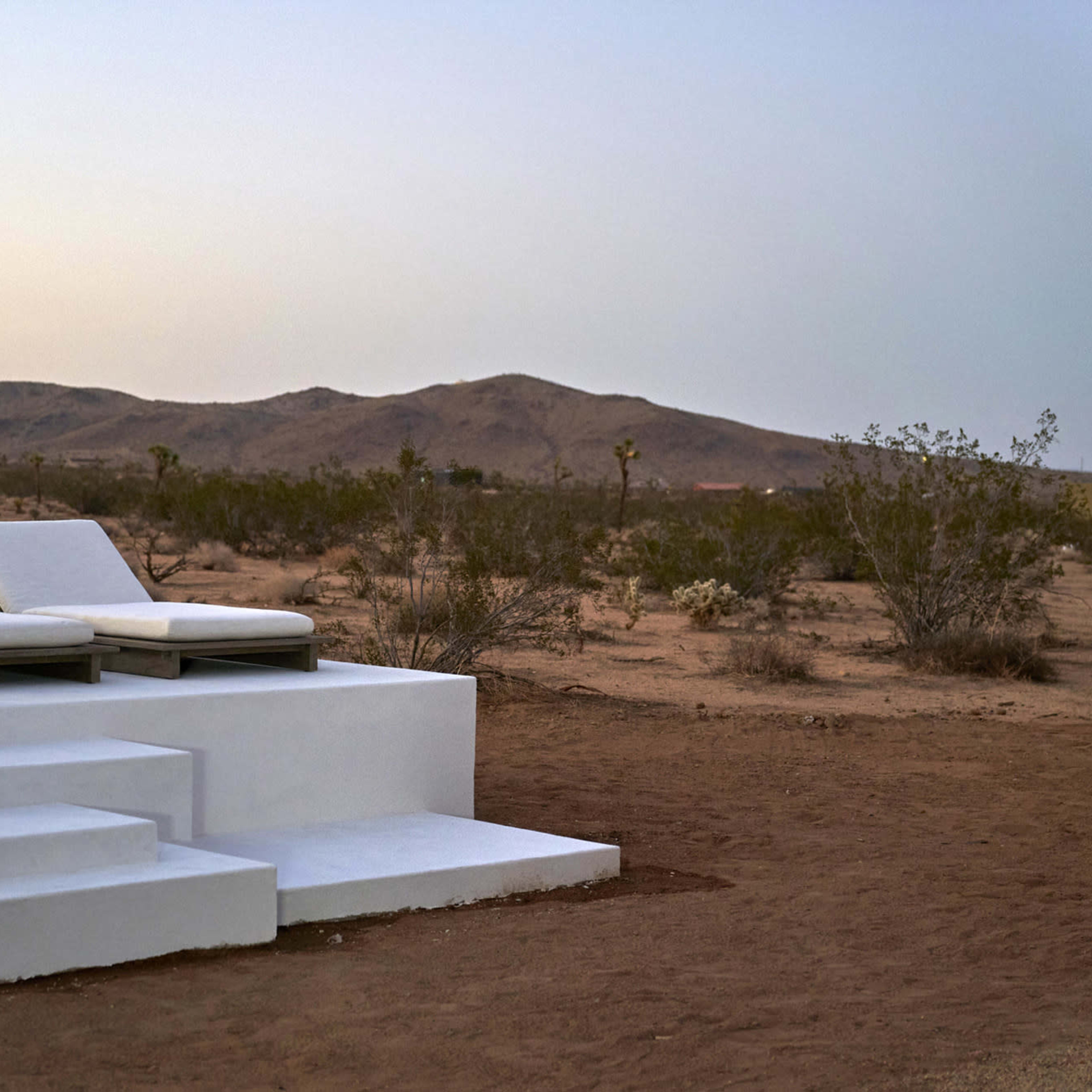 Two white lounge chairs are positioned on a raised platform in a barren desert landscape with rolling hills in the background.