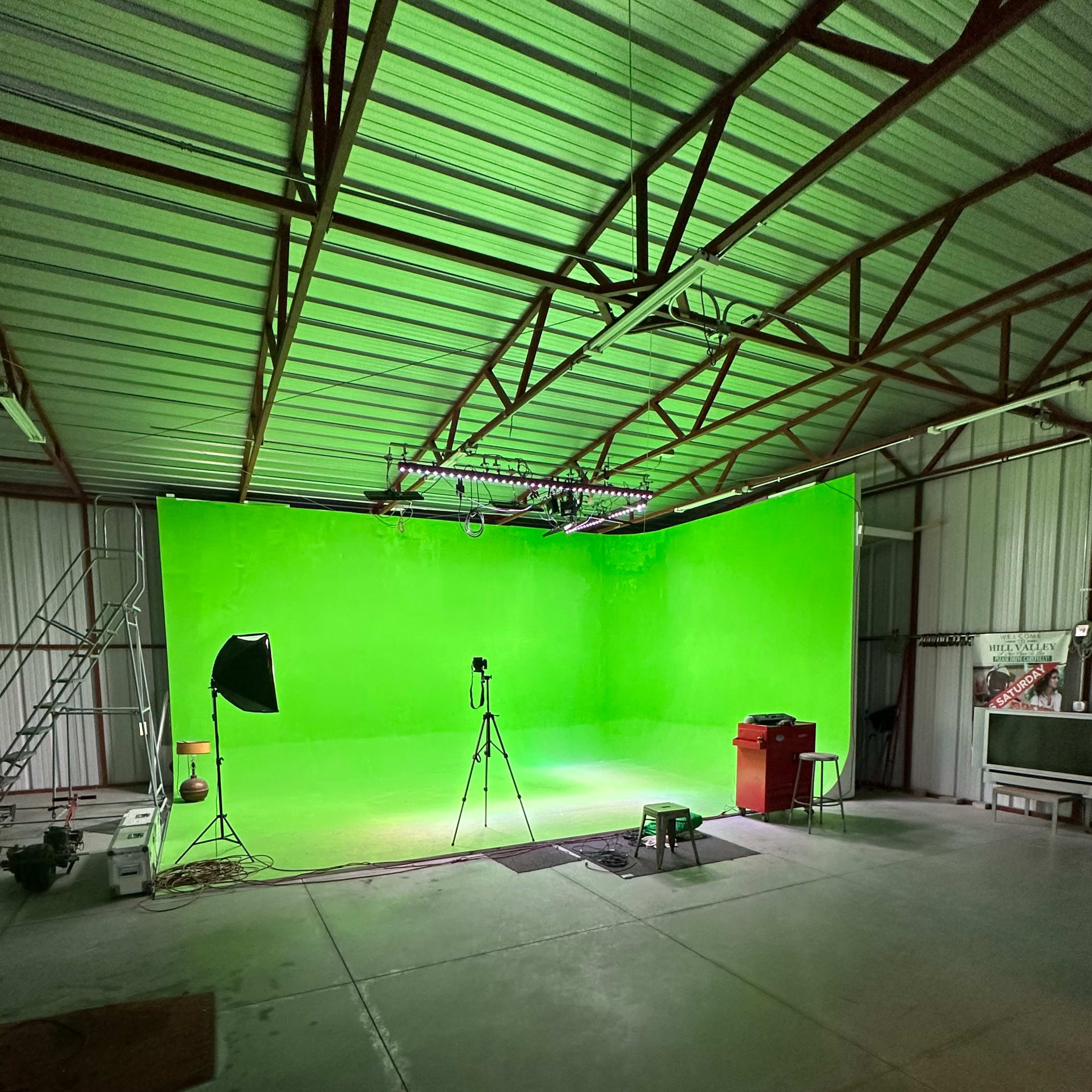 A spacious studio featuring a large green screen, a camera on a tripod, and lighting equipment.