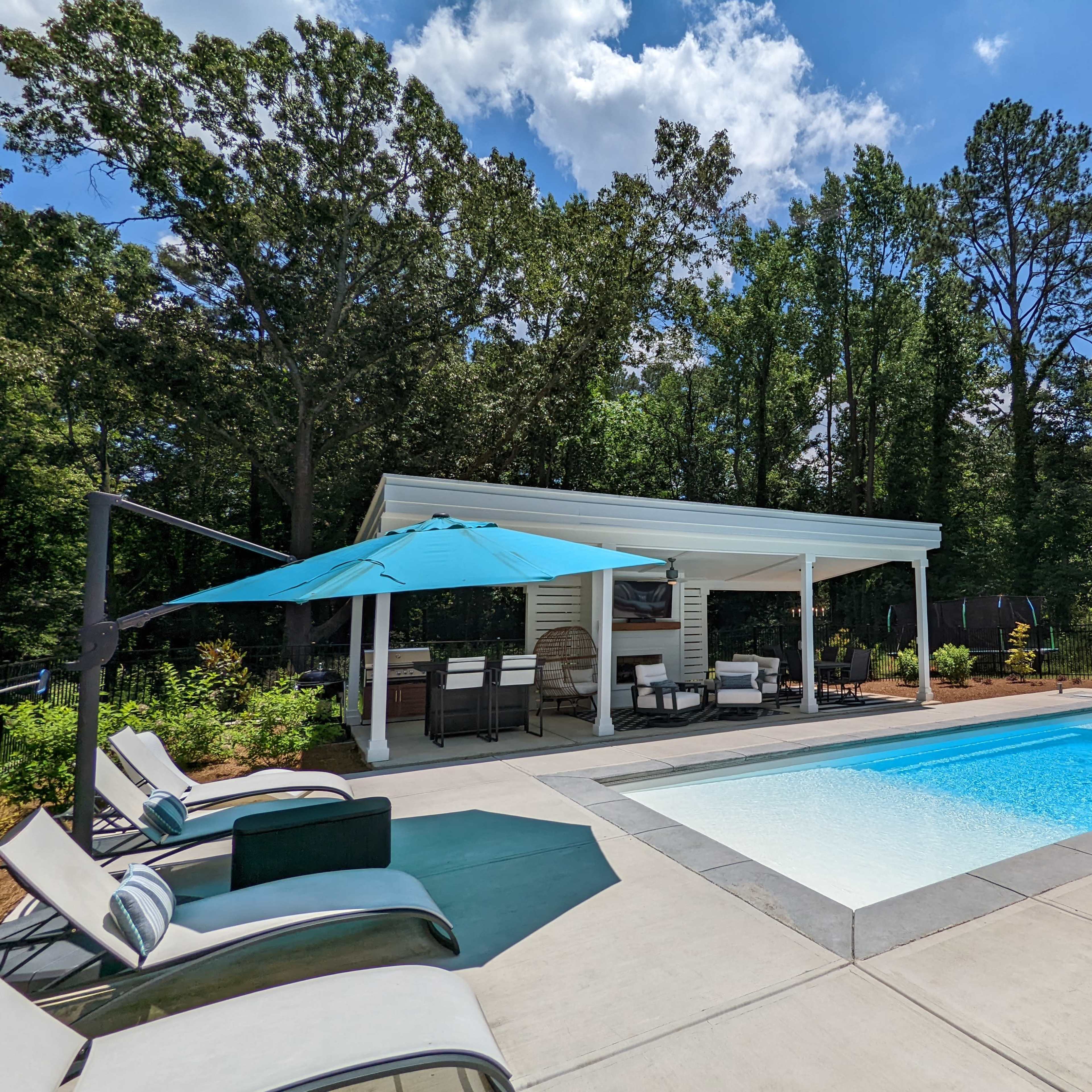 A rectangular swimming pool is surrounded by lounge chairs and a shaded seating area under a white canopy in a lush, green backyard.