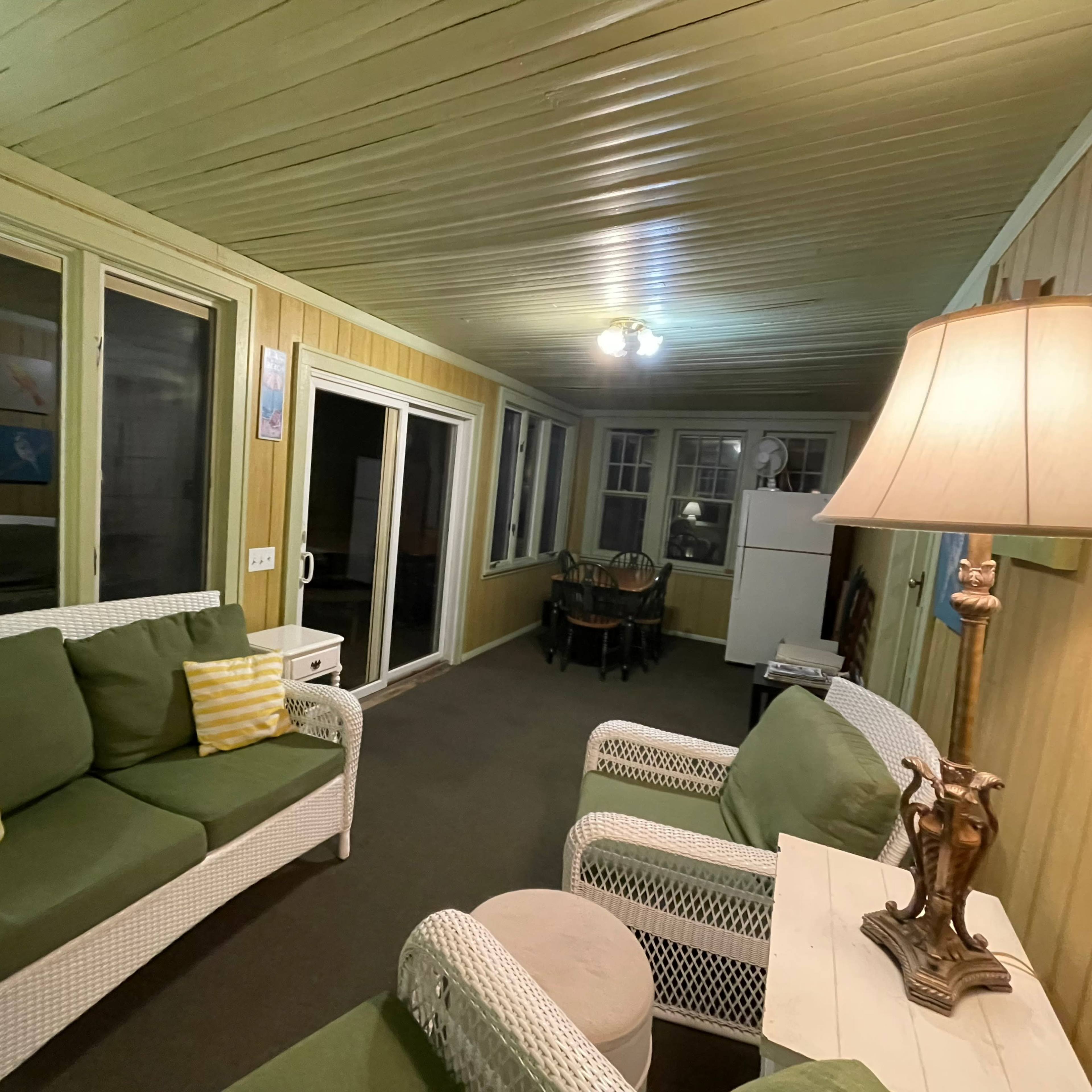 A sunroom with green sofas, a white lamp, and a dining area visible in the background.