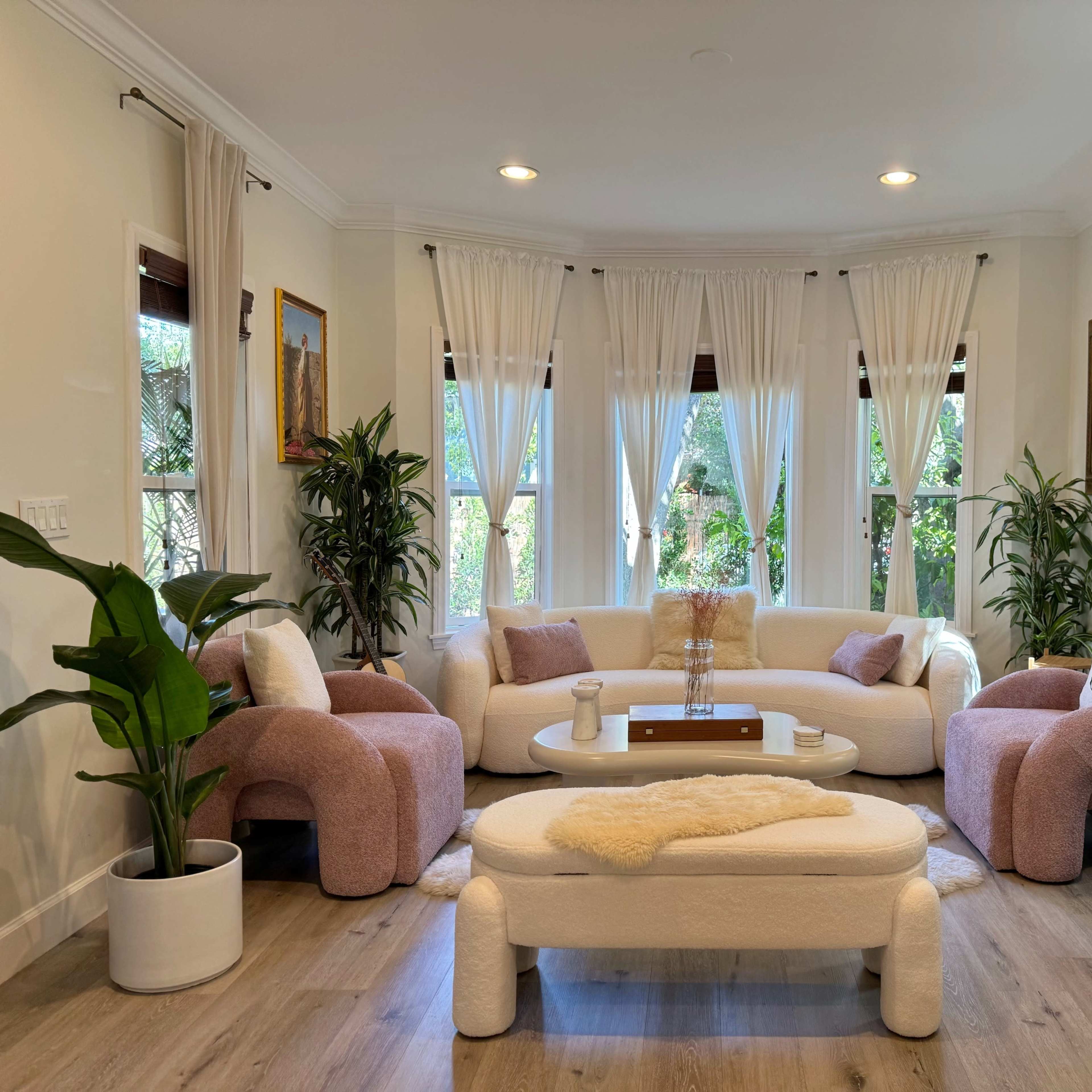 A modern living room with a circular arrangement of soft seating, surrounded by large plants and featuring large windows dressed with sheer curtains.
