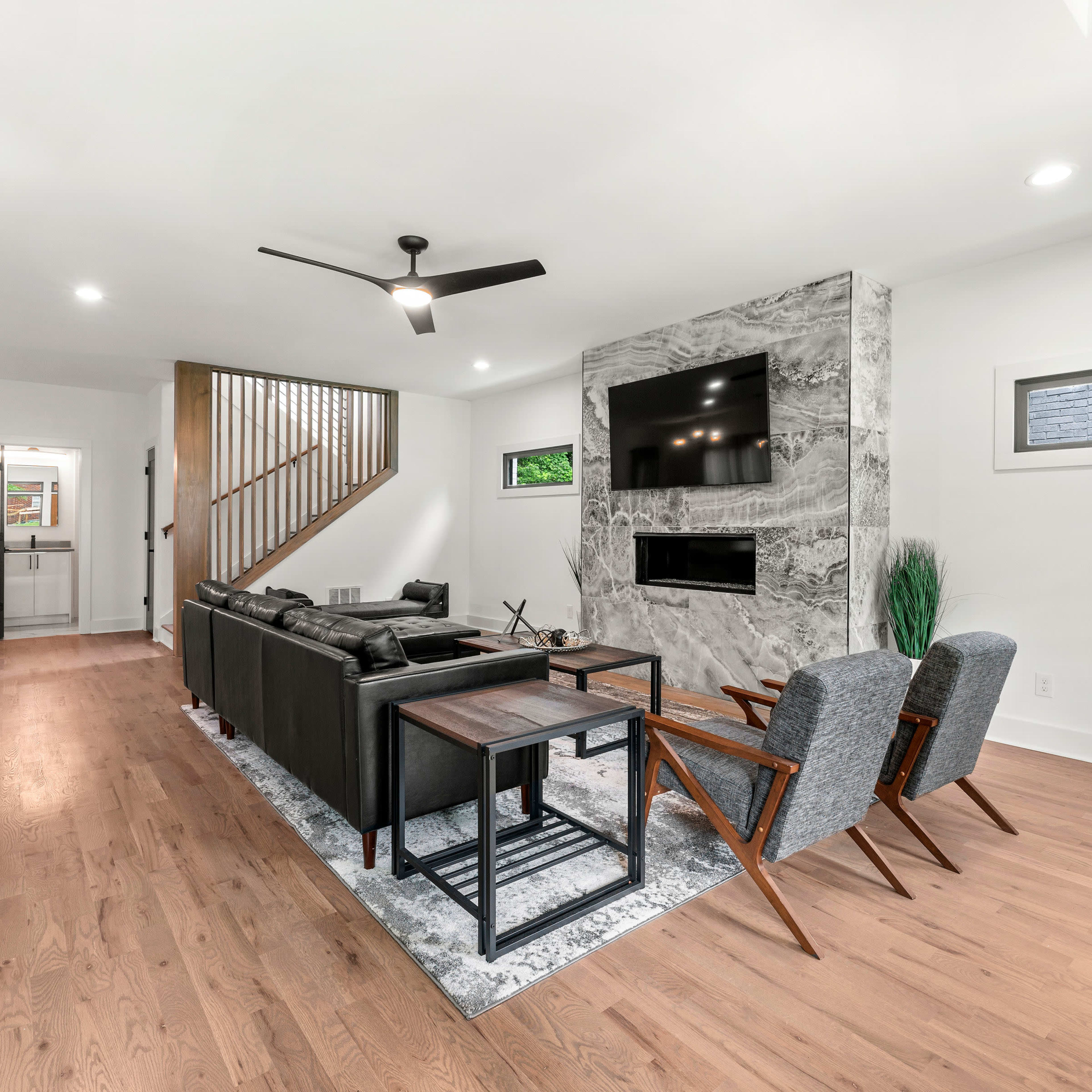 A modern living room featuring a black leather sofa, two gray armchairs, a marble accent wall with a mounted TV, and wooden flooring.