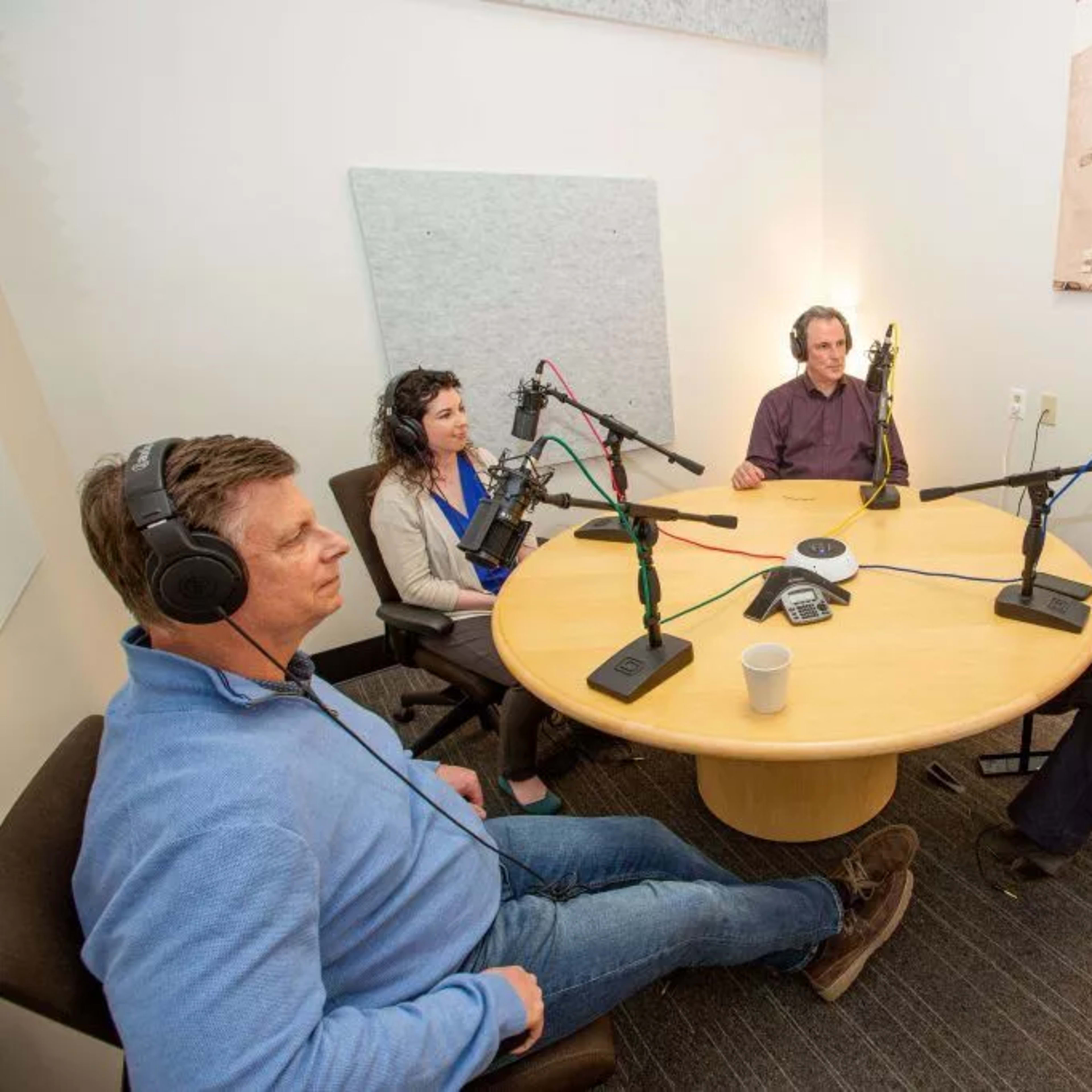 Four people sit around a circular table in a recording studio, each wearing headphones and speaking into microphones.