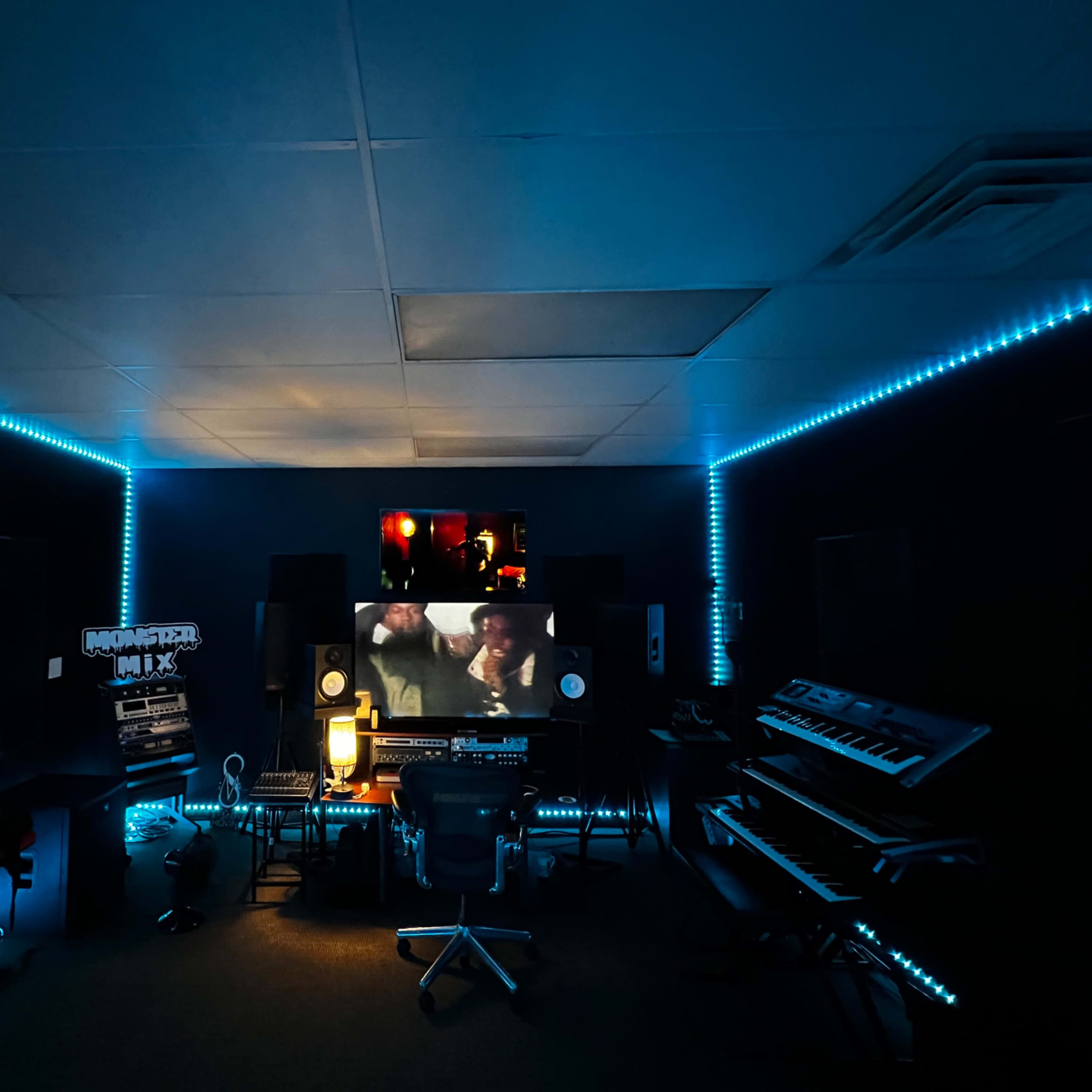 A music studio with illuminated blue LED lights, featuring various musical equipment, a large screen displaying a video, and a central workstation with chairs.
