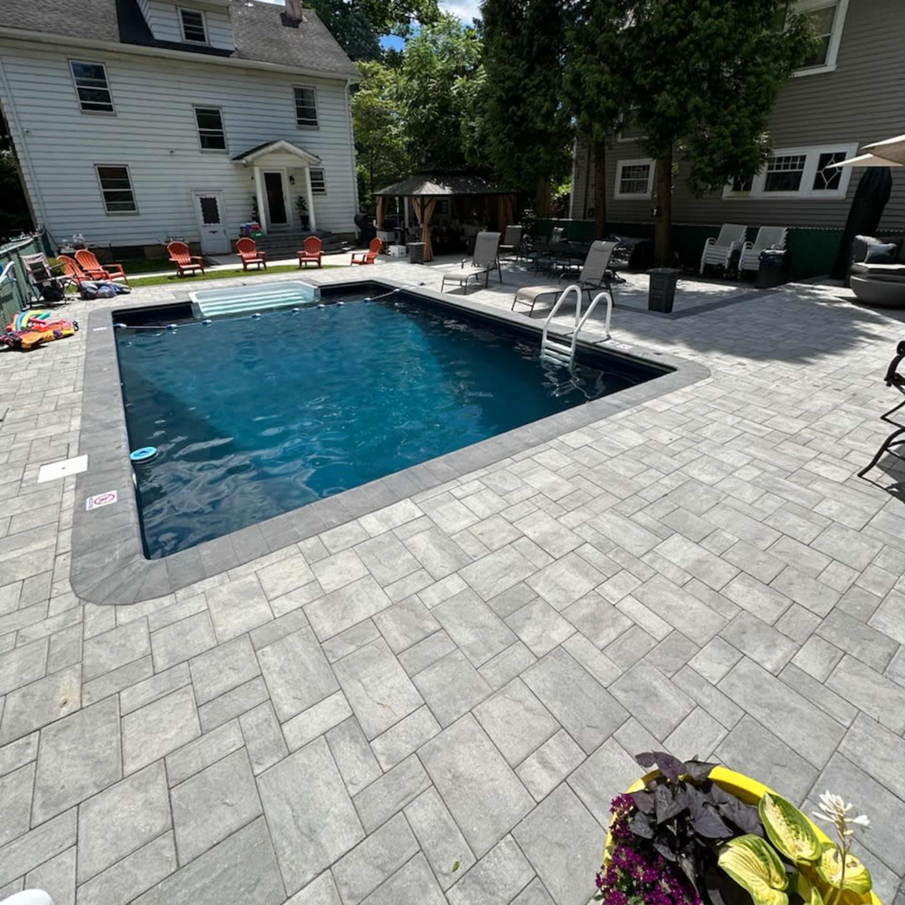 A rectangular swimming pool is surrounded by paved concrete, with lounge chairs and a house visible in the background.