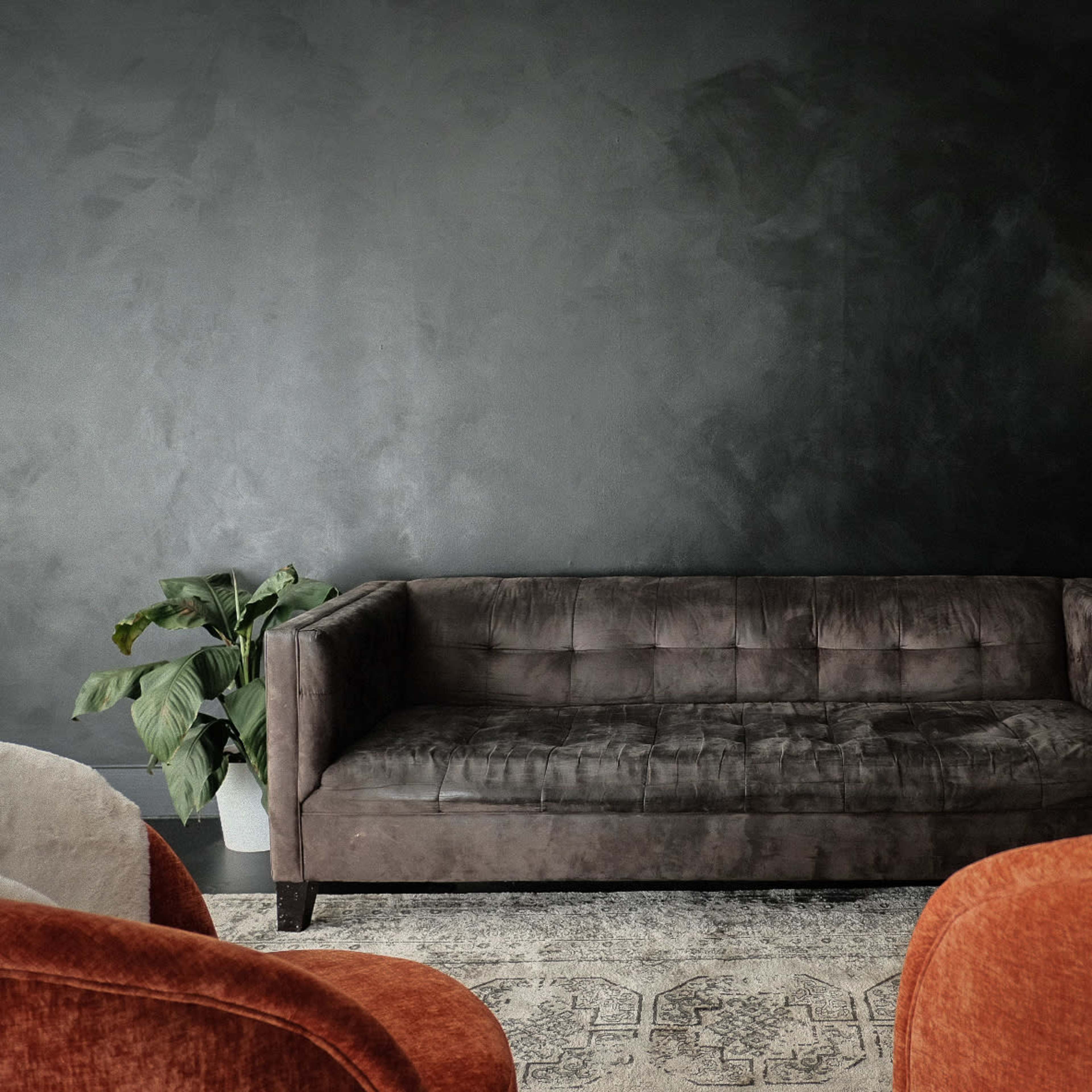 A dark room featuring a brown upholstered sofa, two orange chairs, a floor lamp, and a potted plant.