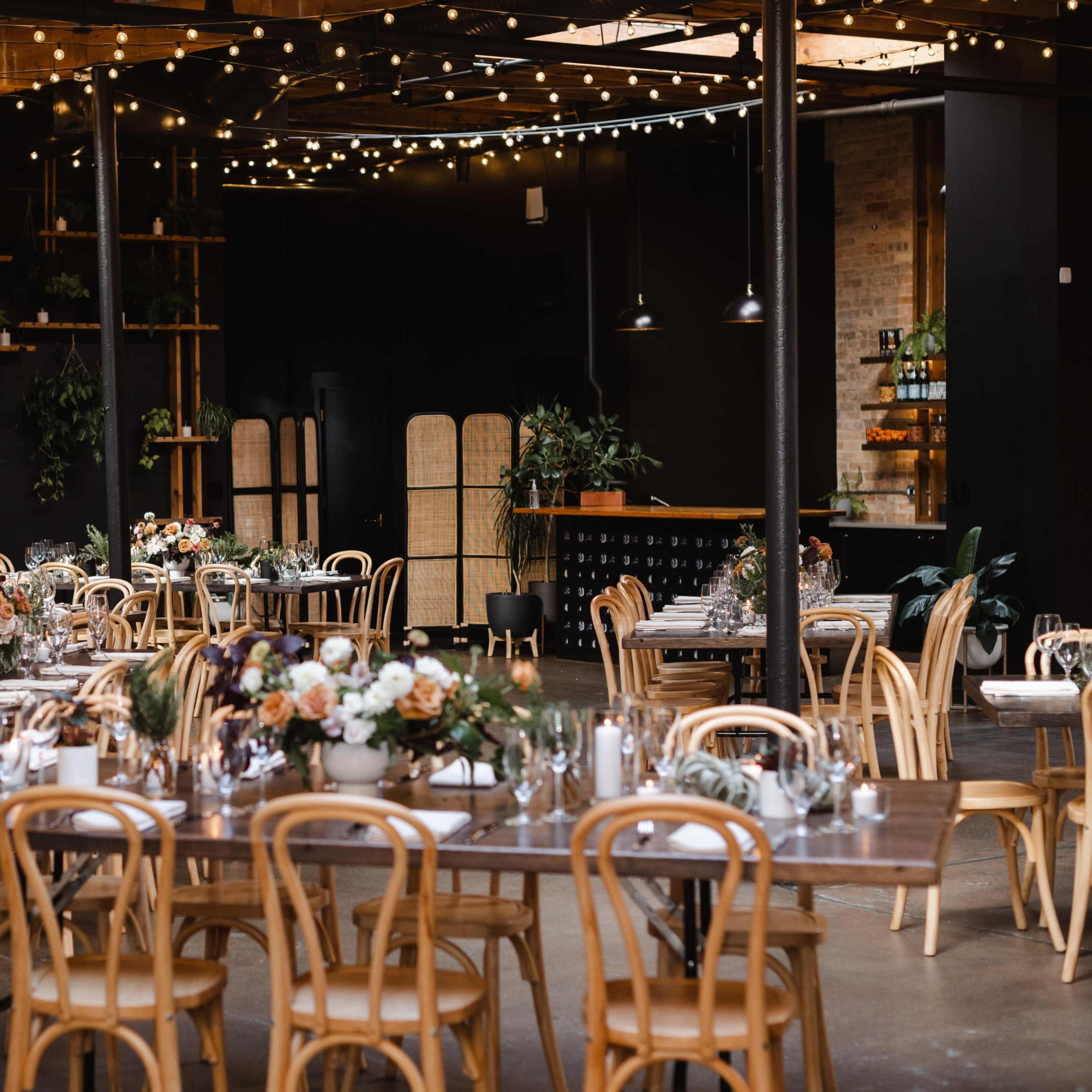 A dimly lit event space features long wooden tables elegantly arranged with floral centerpieces and surrounded by bentwood chairs, with string lights overhead and greenery adorning the walls.