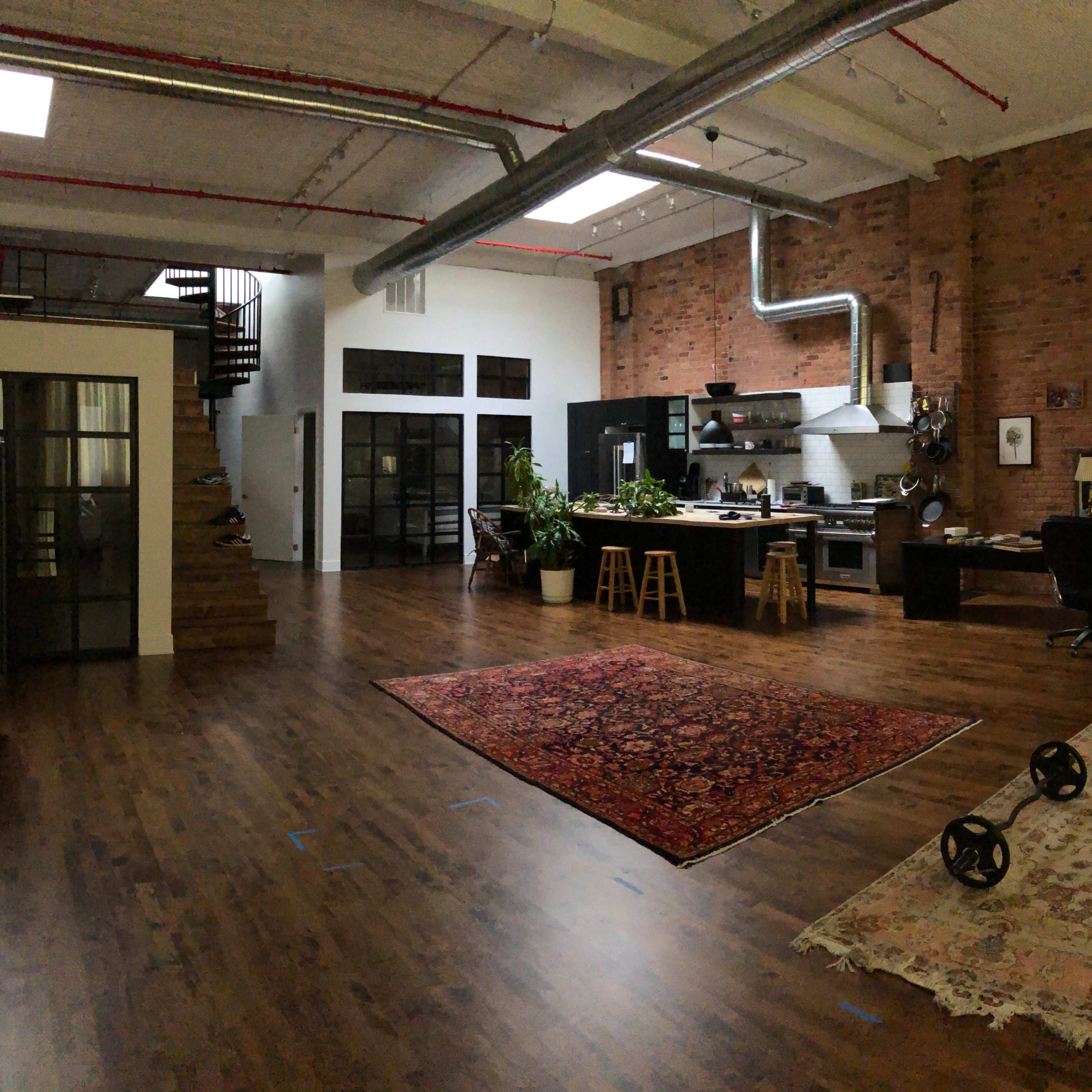 A spacious loft-style interior with exposed brick walls, a kitchen area, a dining space, and a living room featuring rugs and workout equipment.