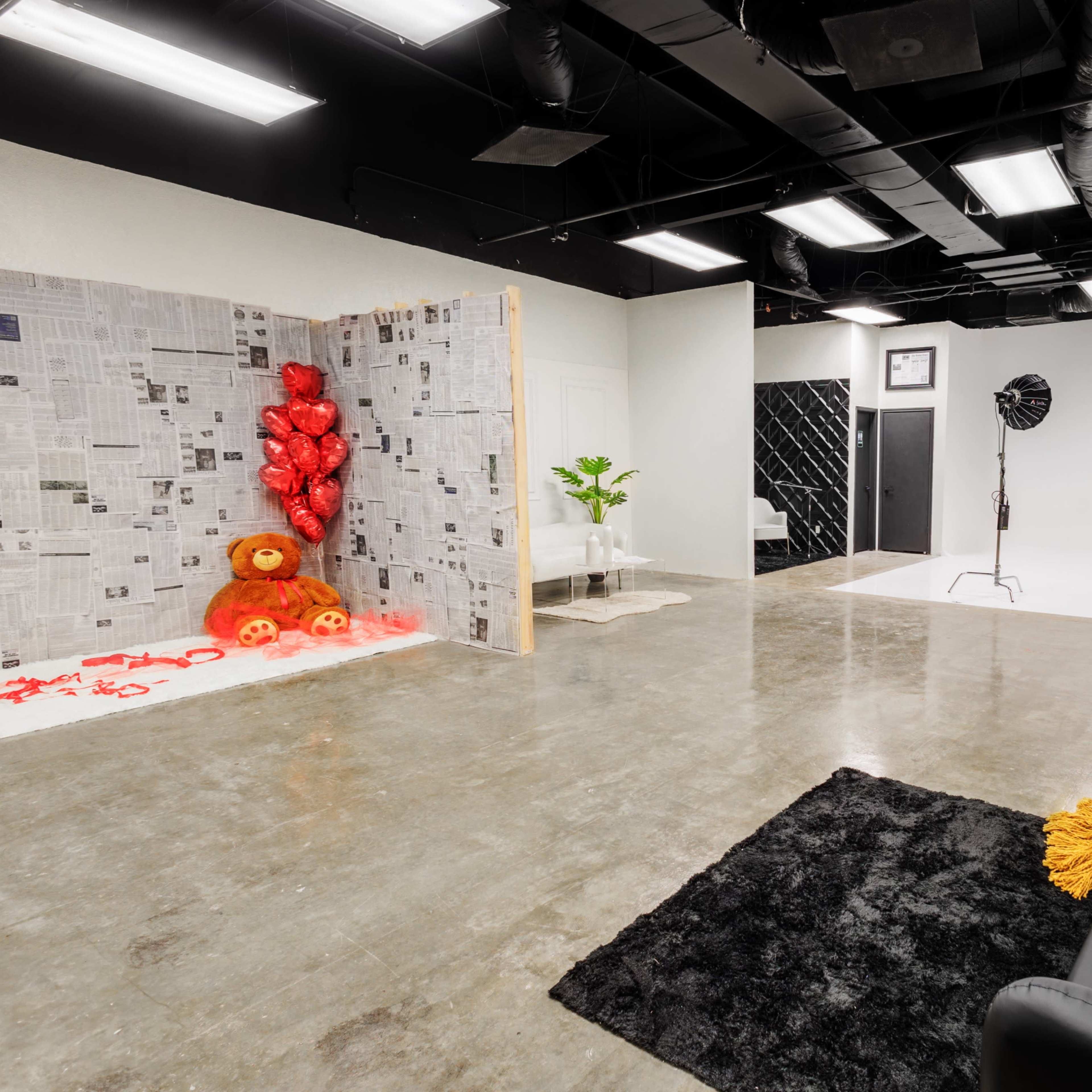 A photography studio featuring a black sofa, a section with newspaper-covered walls and red heart balloons, and a backdrop area with lighting equipment.