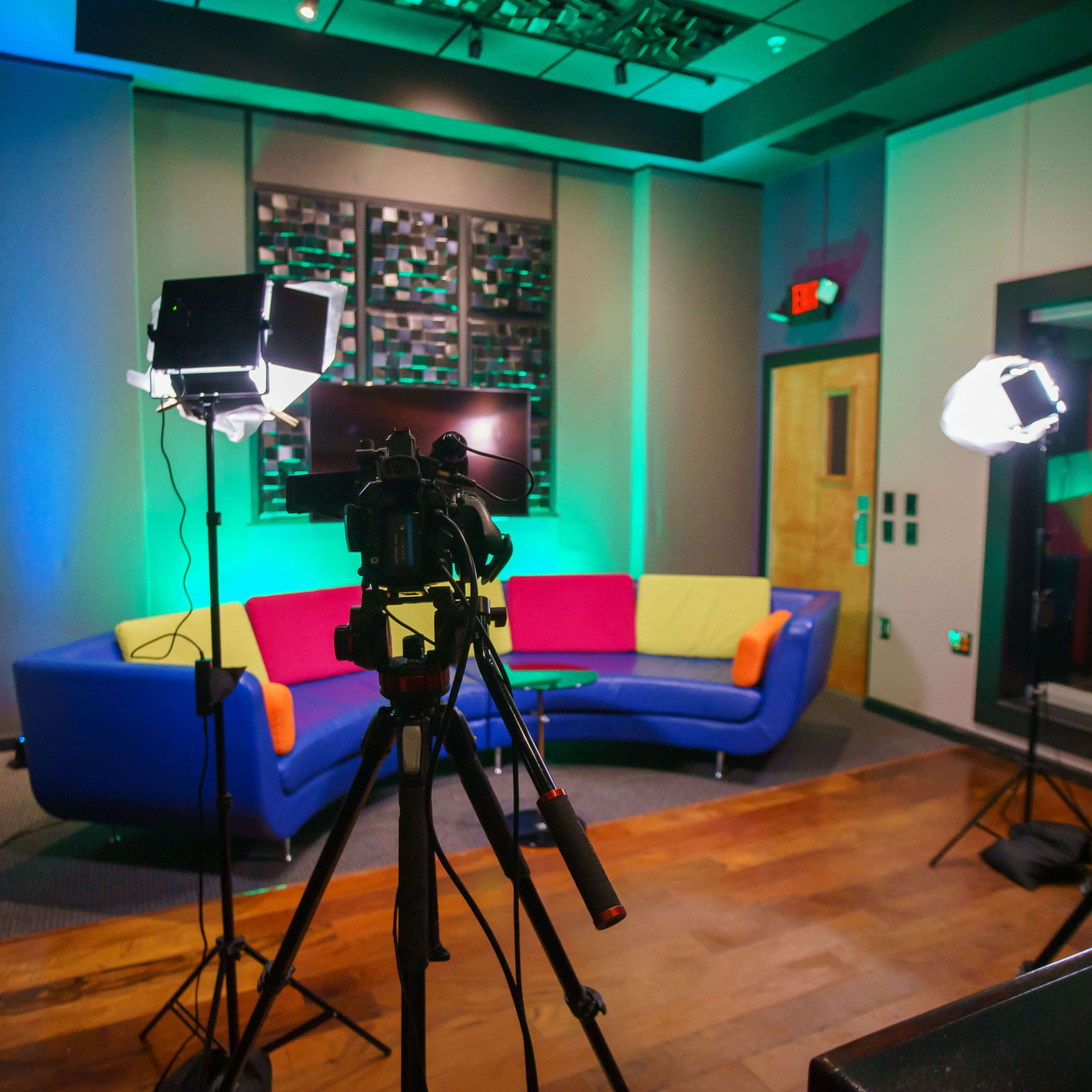 A brightly lit studio setup featuring a colorful couch, two studio lights, and cameras positioned on tripods.