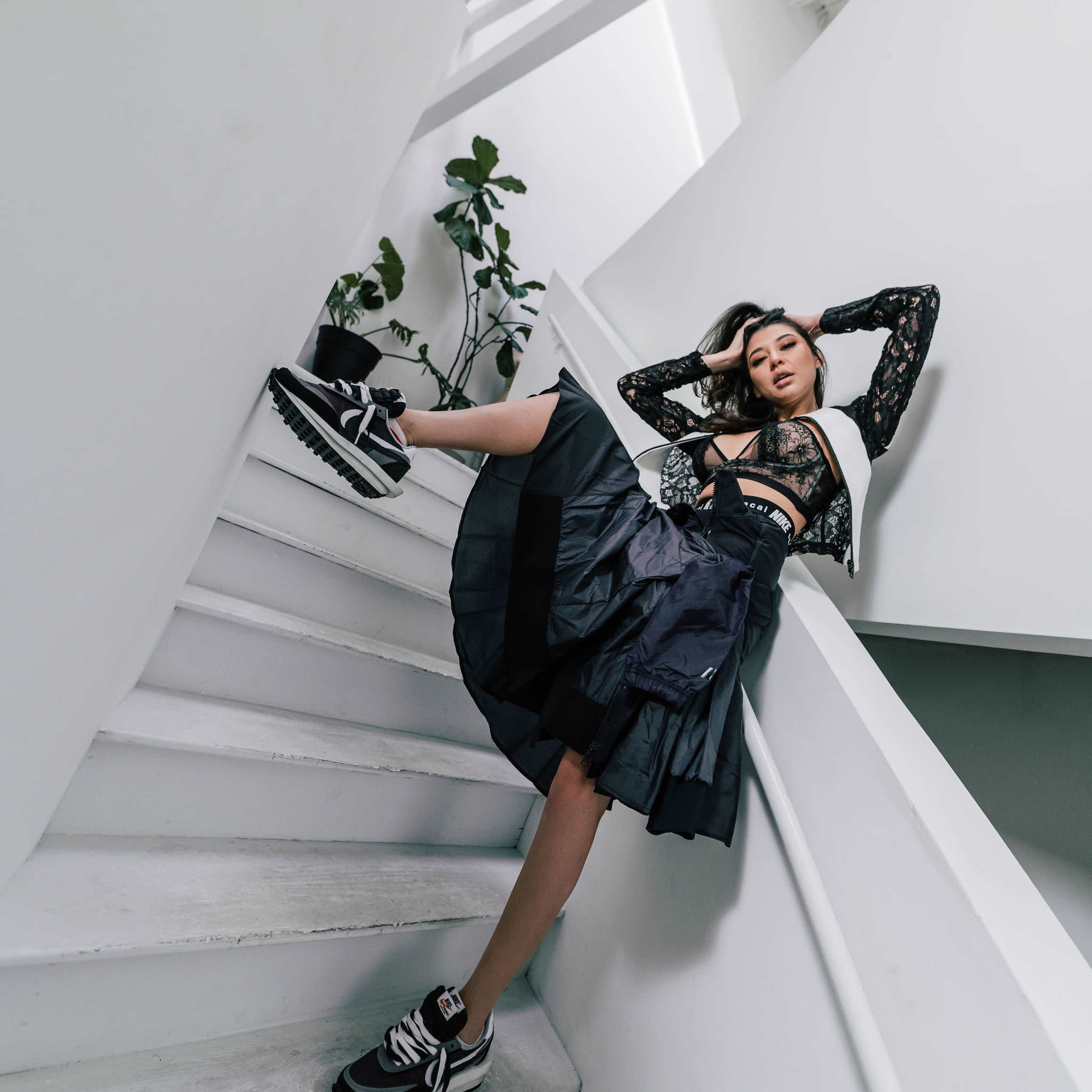 A person poses on a staircase, wearing a black lace top and a voluminous black skirt, while leaning against the wall.