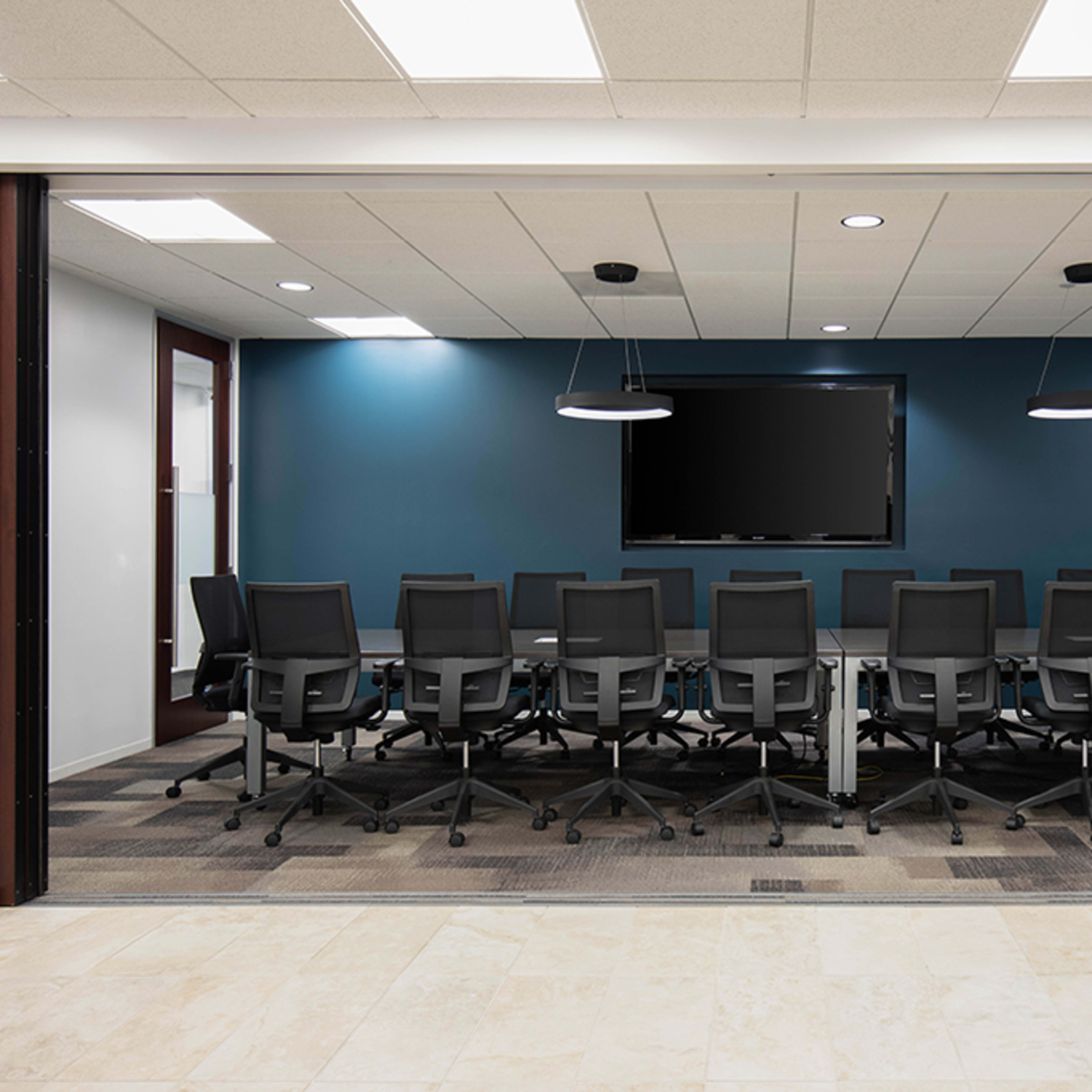 A modern conference room features a long table surrounded by ergonomic black office chairs, with a large screen mounted on a teal wall.