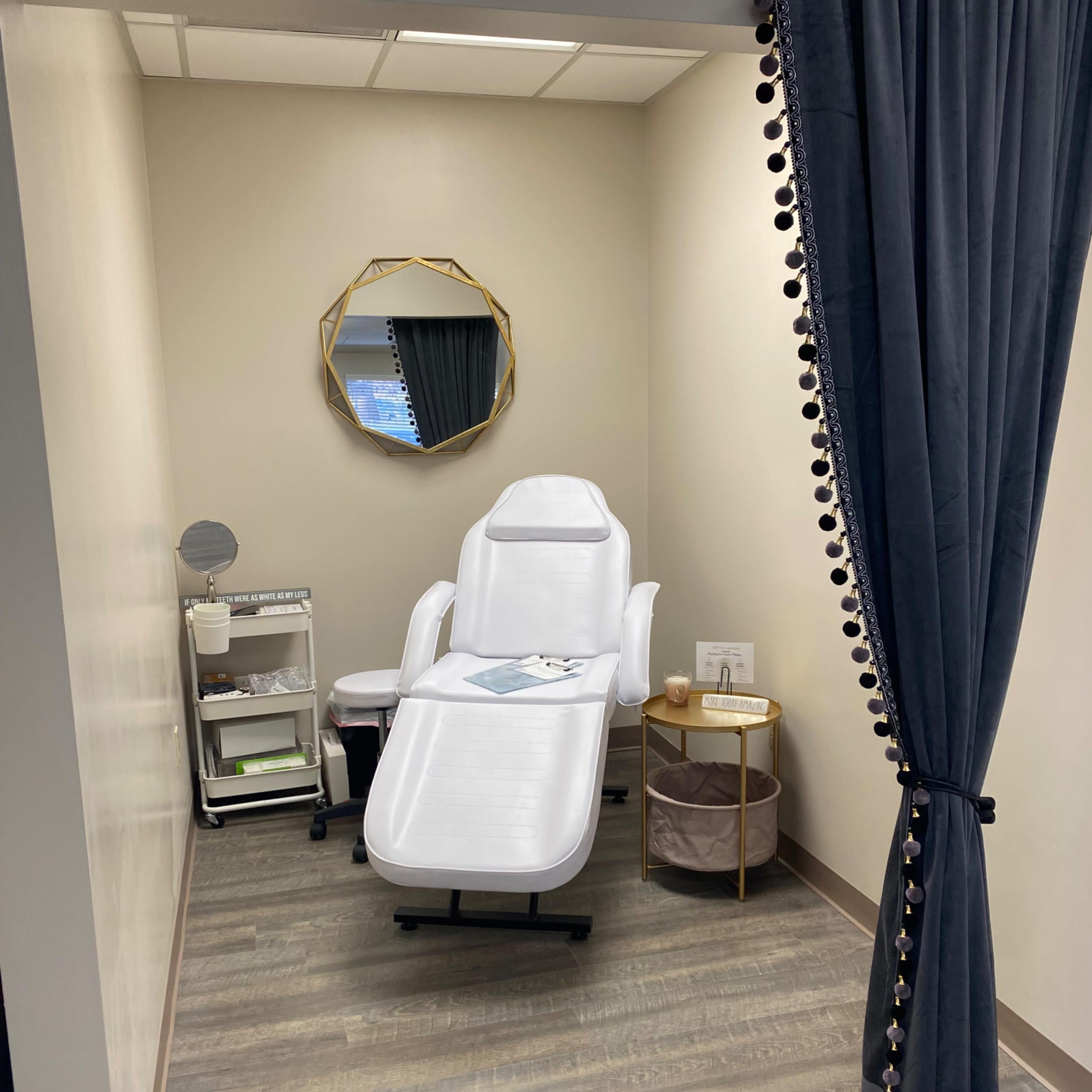 A small treatment room with a white reclining chair, a cart with supplies, and a curtain separating it from the rest of the space.