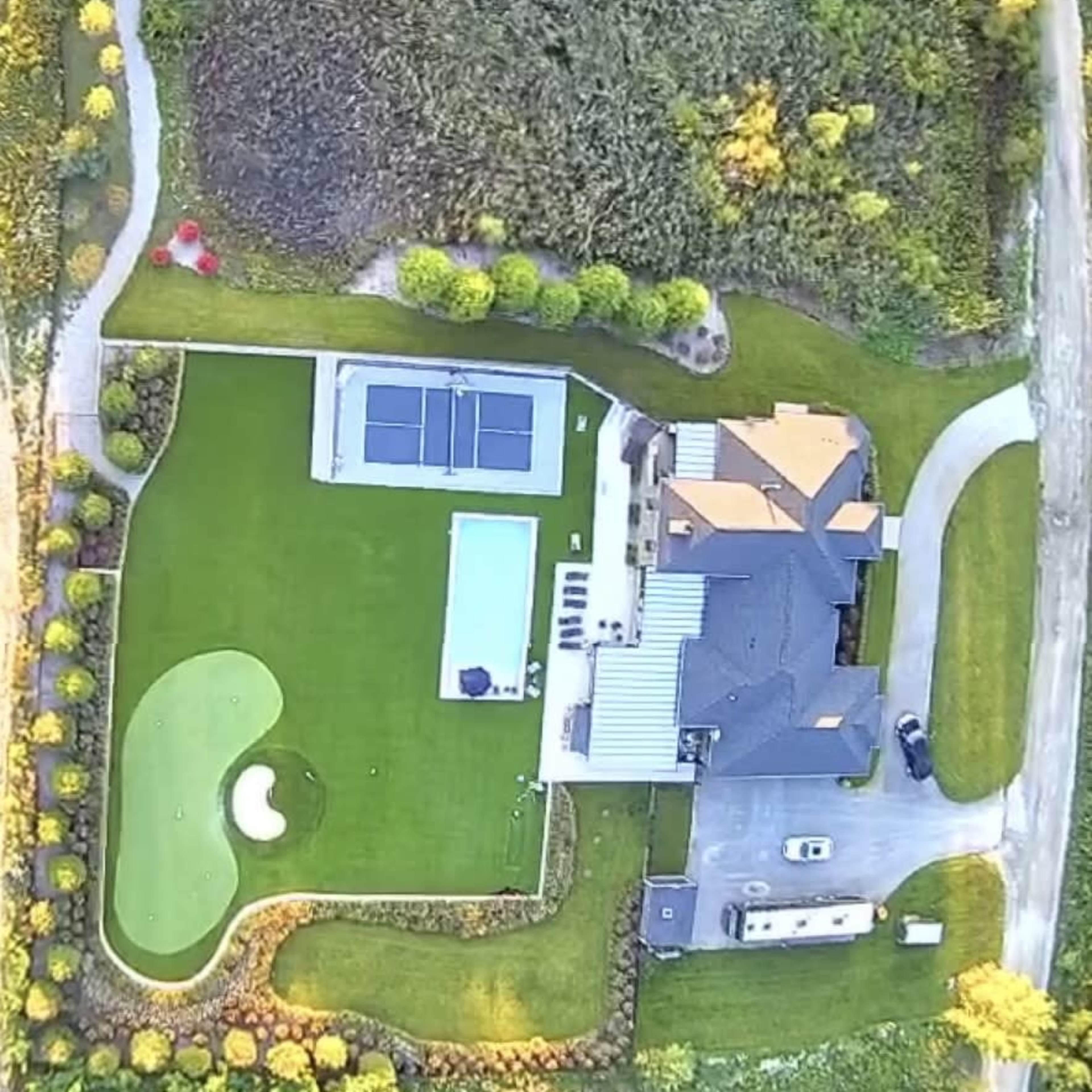 An aerial view of a landscaped property featuring a home, a swimming pool, a tennis court, and a putting green on a large lawn.