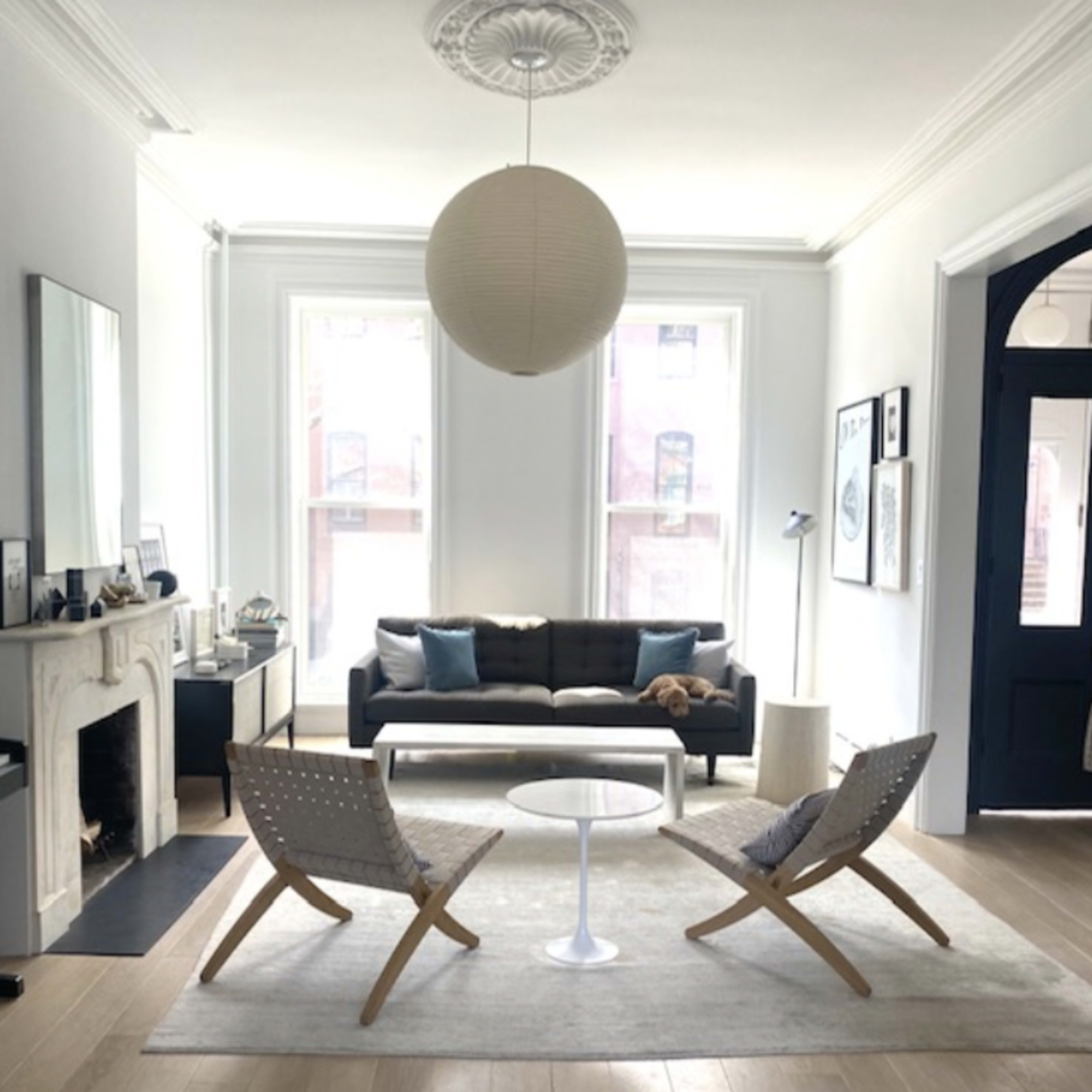 A modern living room with a black sofa, a white circular coffee table, two woven chairs, and large windows allowing natural light.