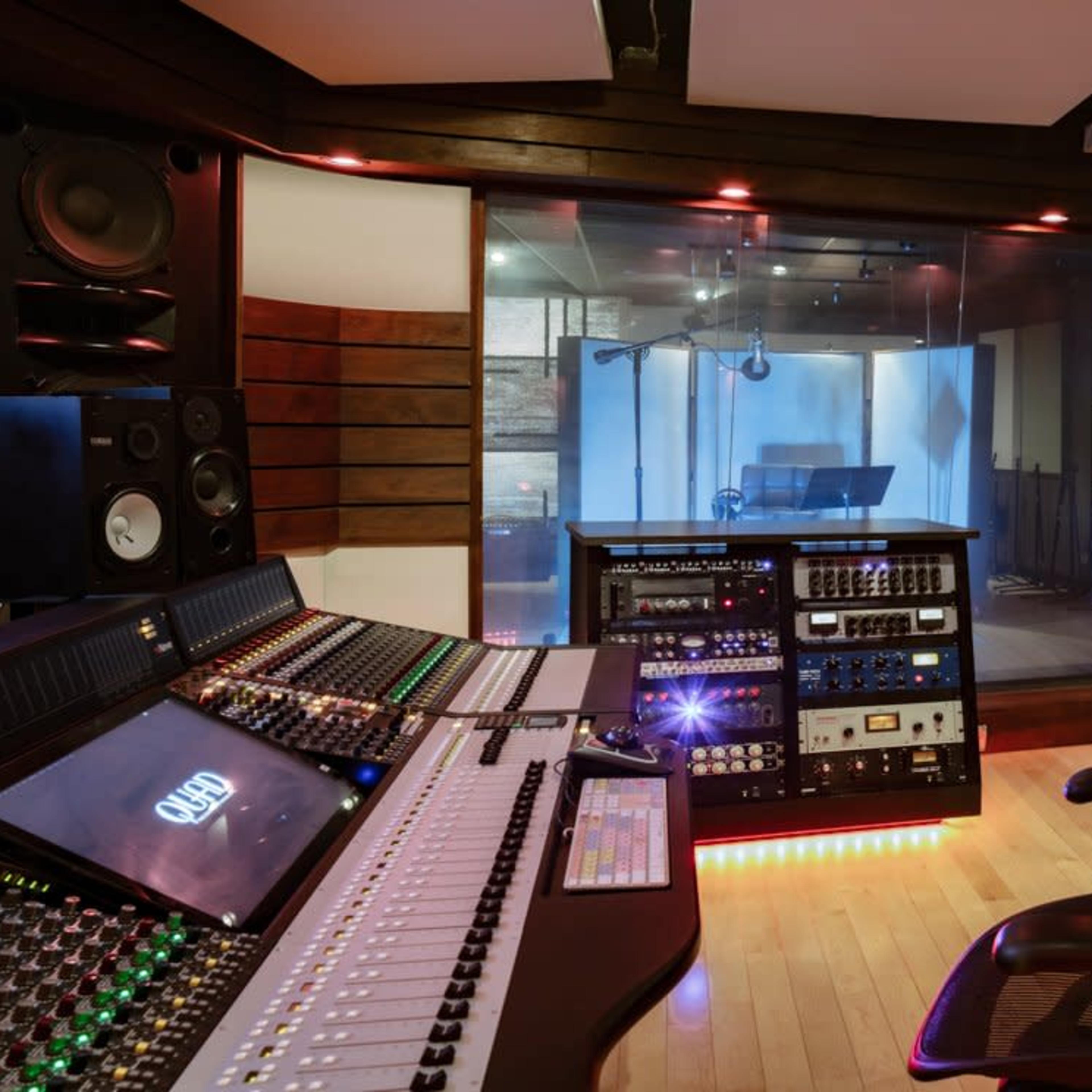 A modern music studio featuring a mixing console, a computer monitor, and a glass-walled recording booth.