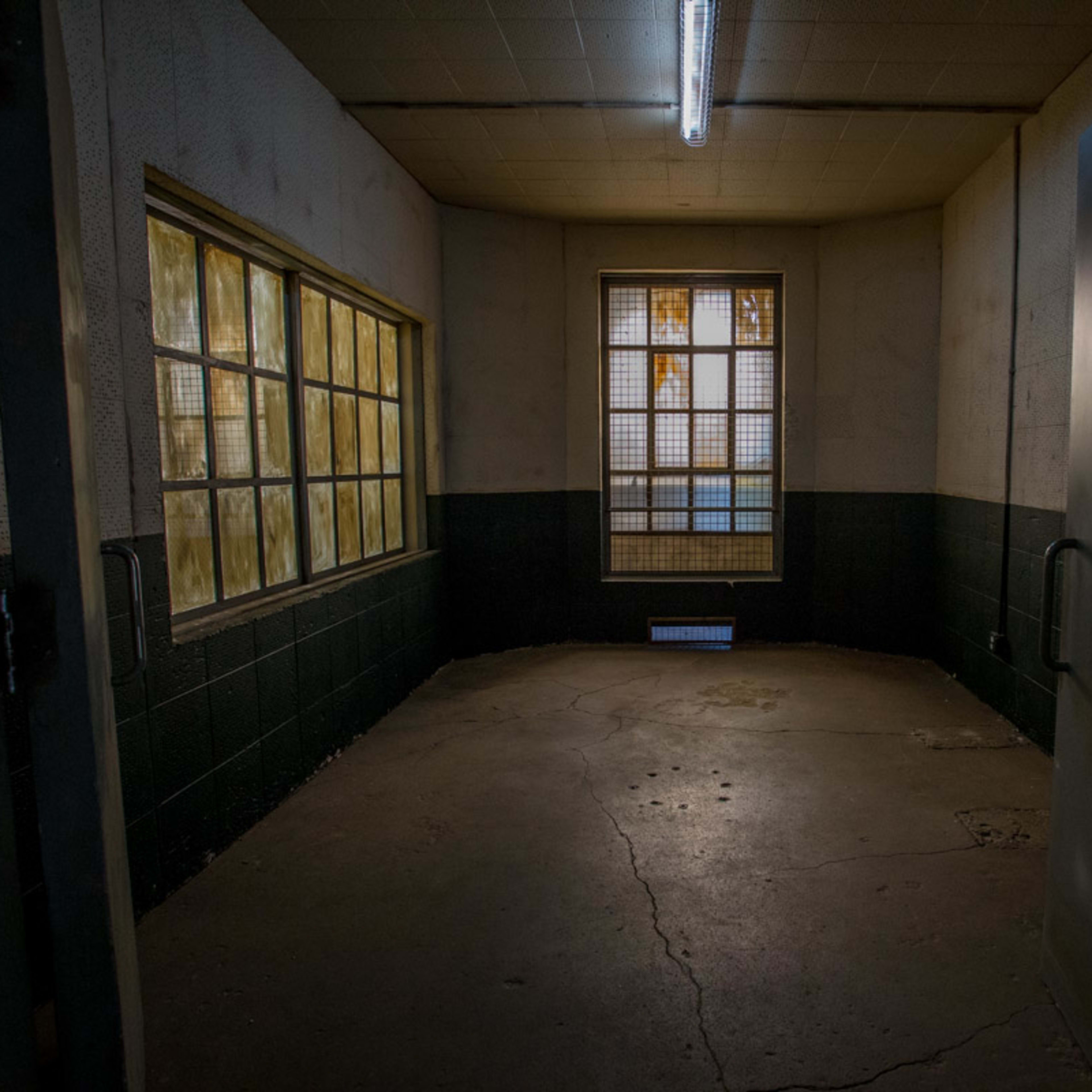 A dimly lit, empty room with concrete floors and two barred windows on one wall.