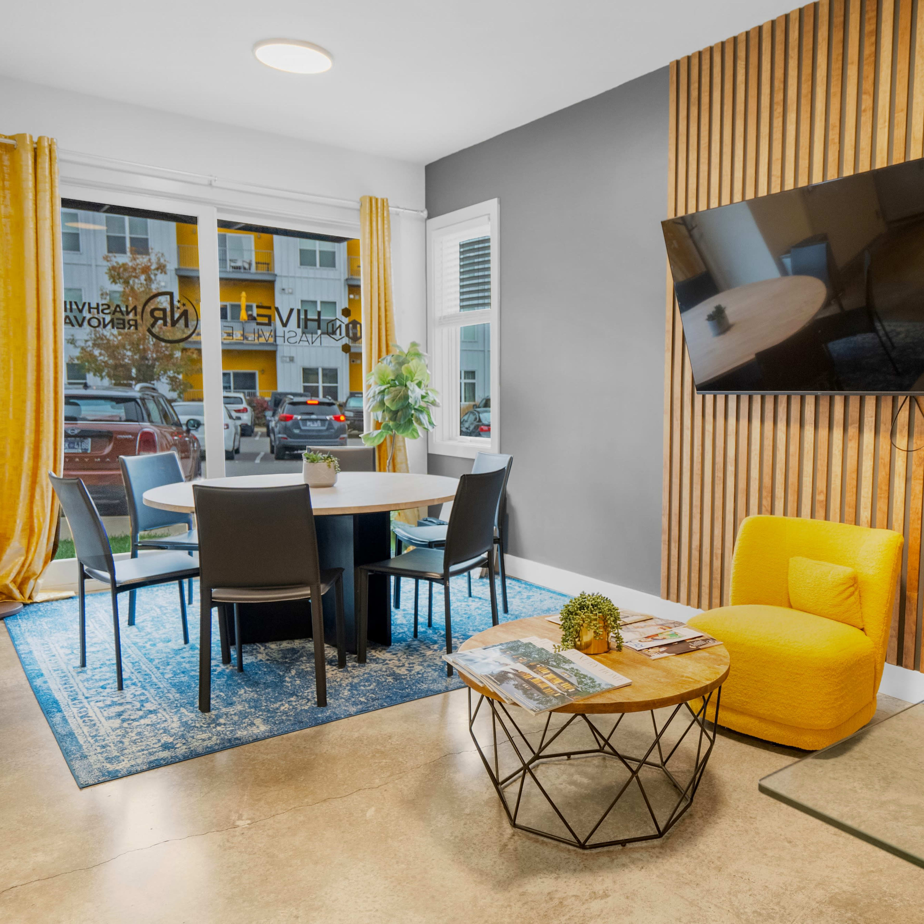A modern sitting area with a round table and chairs, a yellow accent chair, a wooden wall, and a television mounted on the wall.
