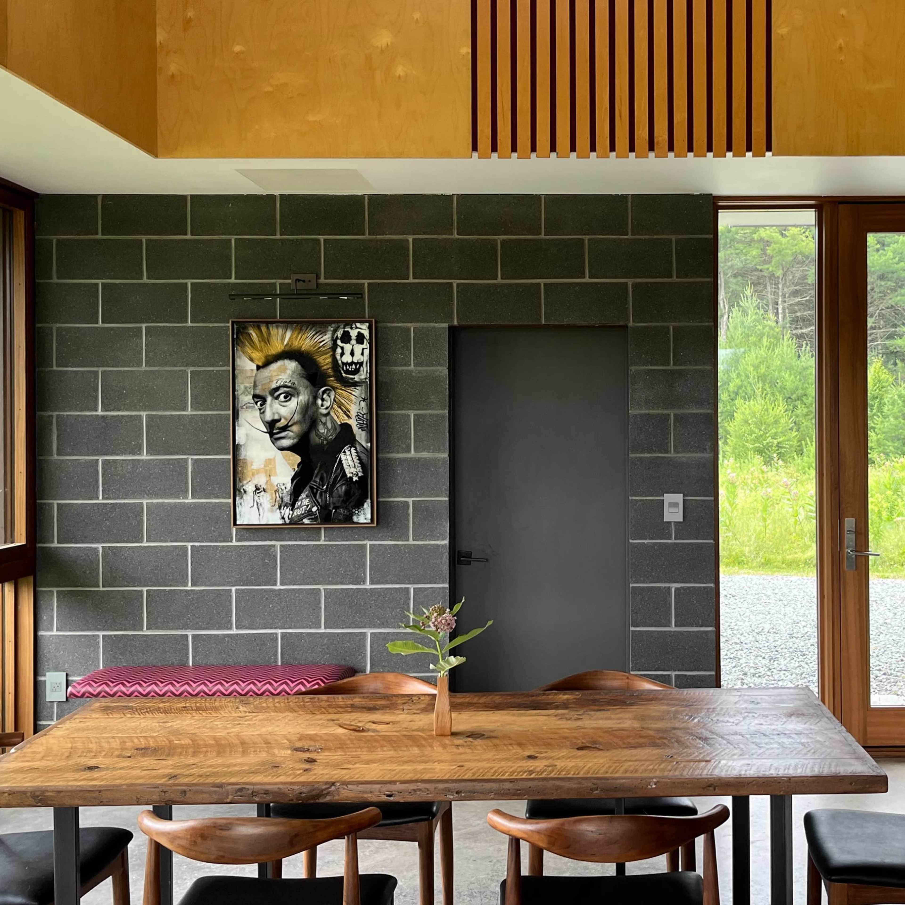 A modern dining area with a wooden table surrounded by dark chairs, a piece of wall art, and large windows overlooking a green landscape.