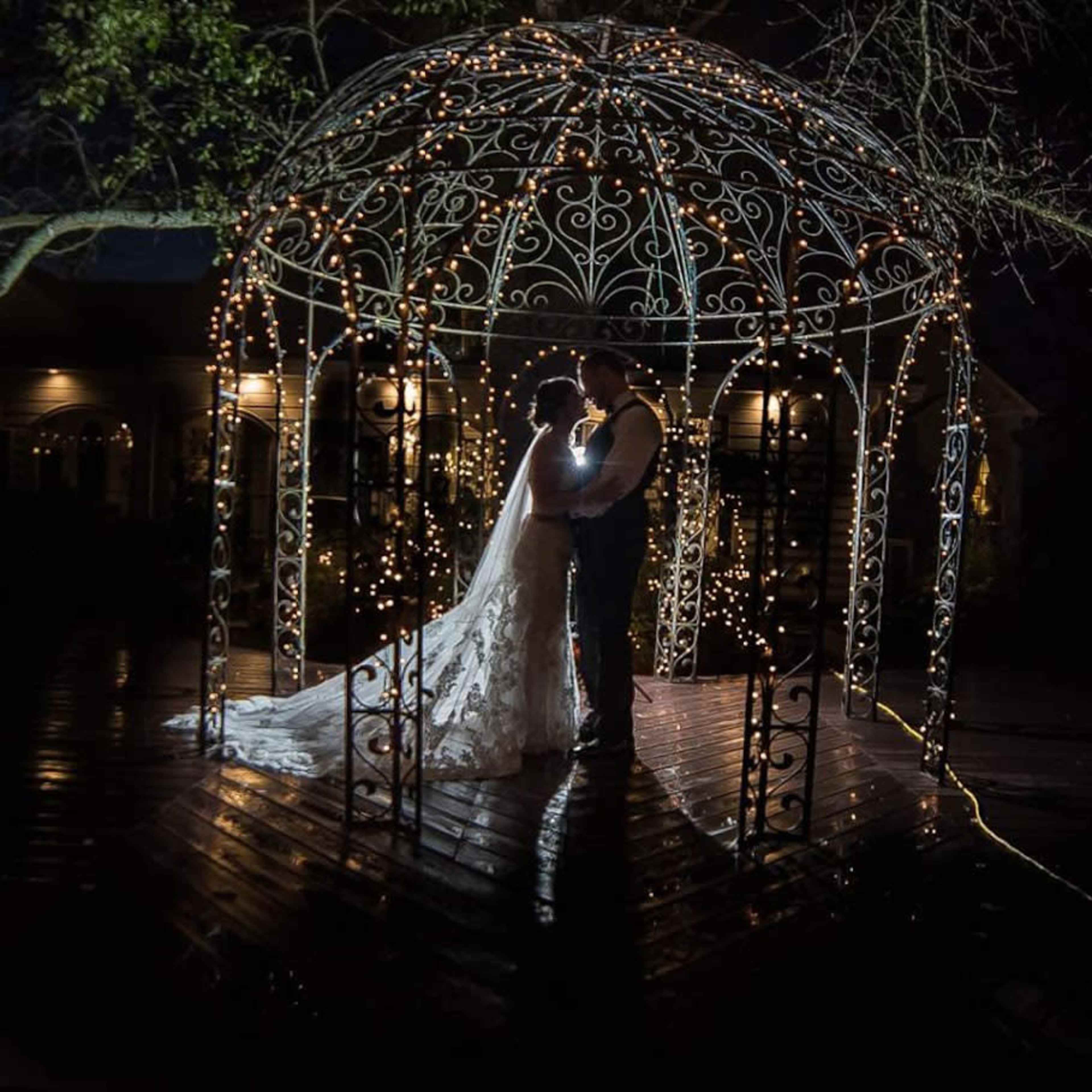 A couple stands under a lit gazebo at night, surrounded by a reflective wooden deck and trees.