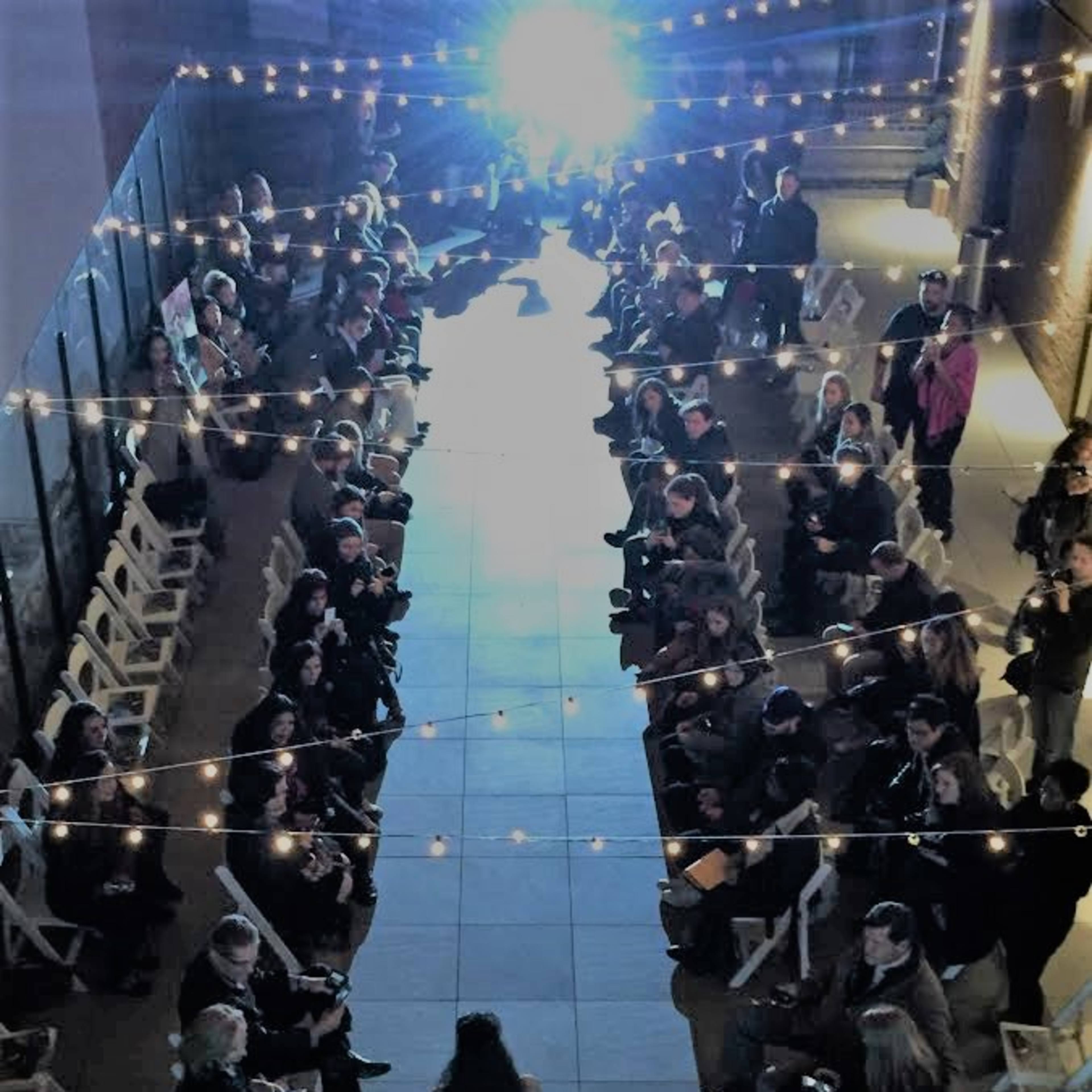 A model walks down a runway illuminated by overhead lights, flanked by seated guests on either side.