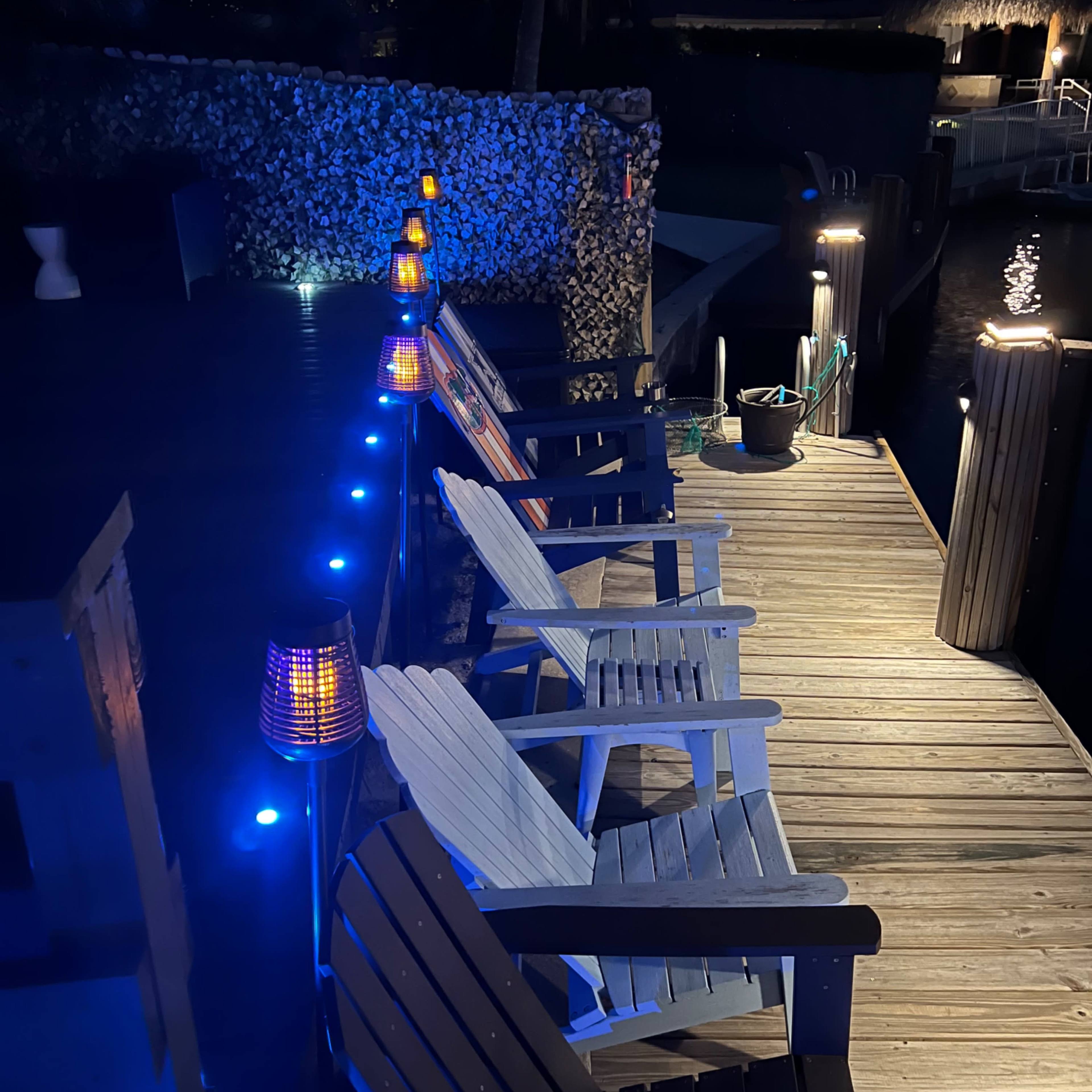 A dock area is illuminated by blue lights and features several white adirondack chairs arranged along a wooden pathway.