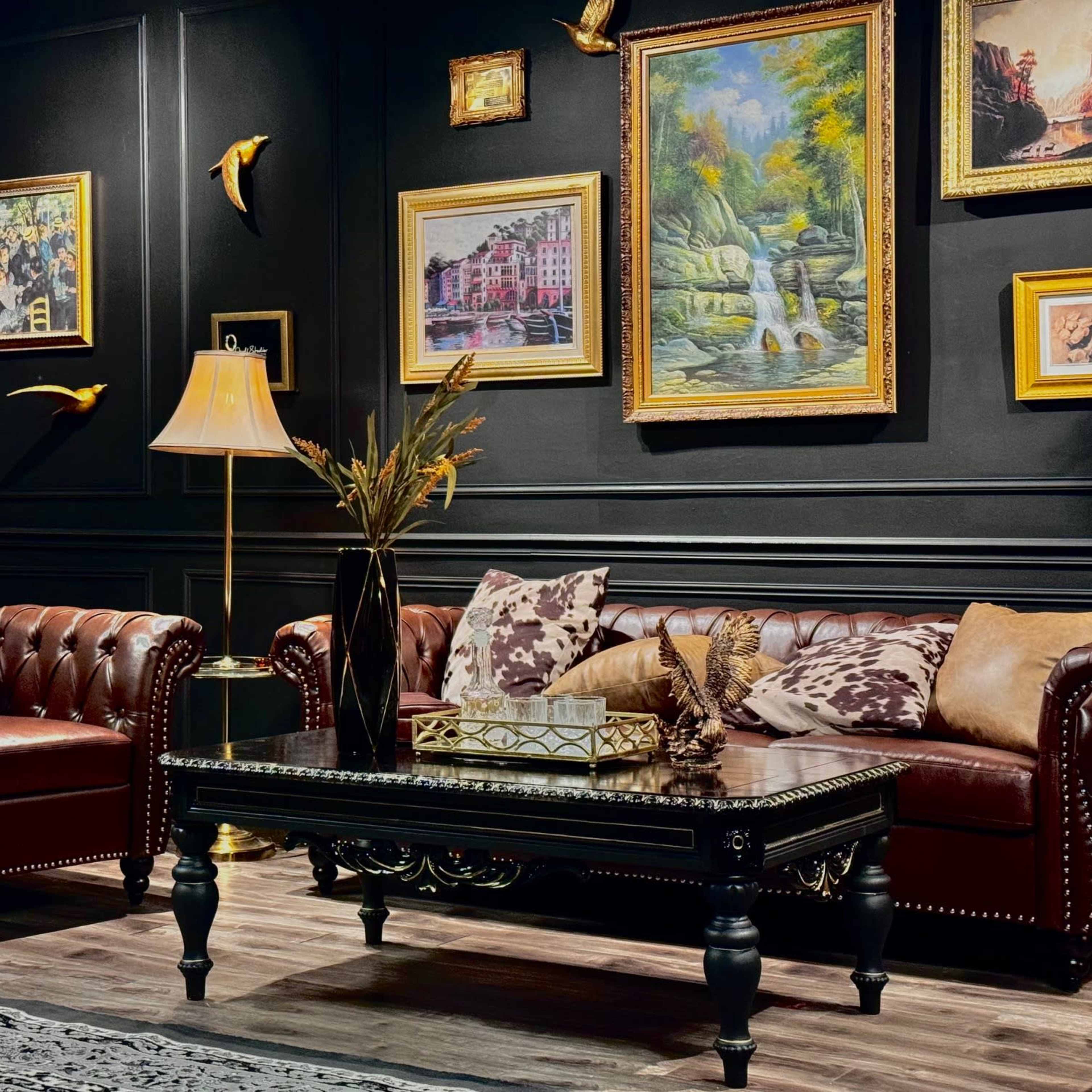 A living room with a dark wall adorned with various framed paintings, featuring a leather chesterfield sofa, an armchair, a decorative coffee table, and lamps.