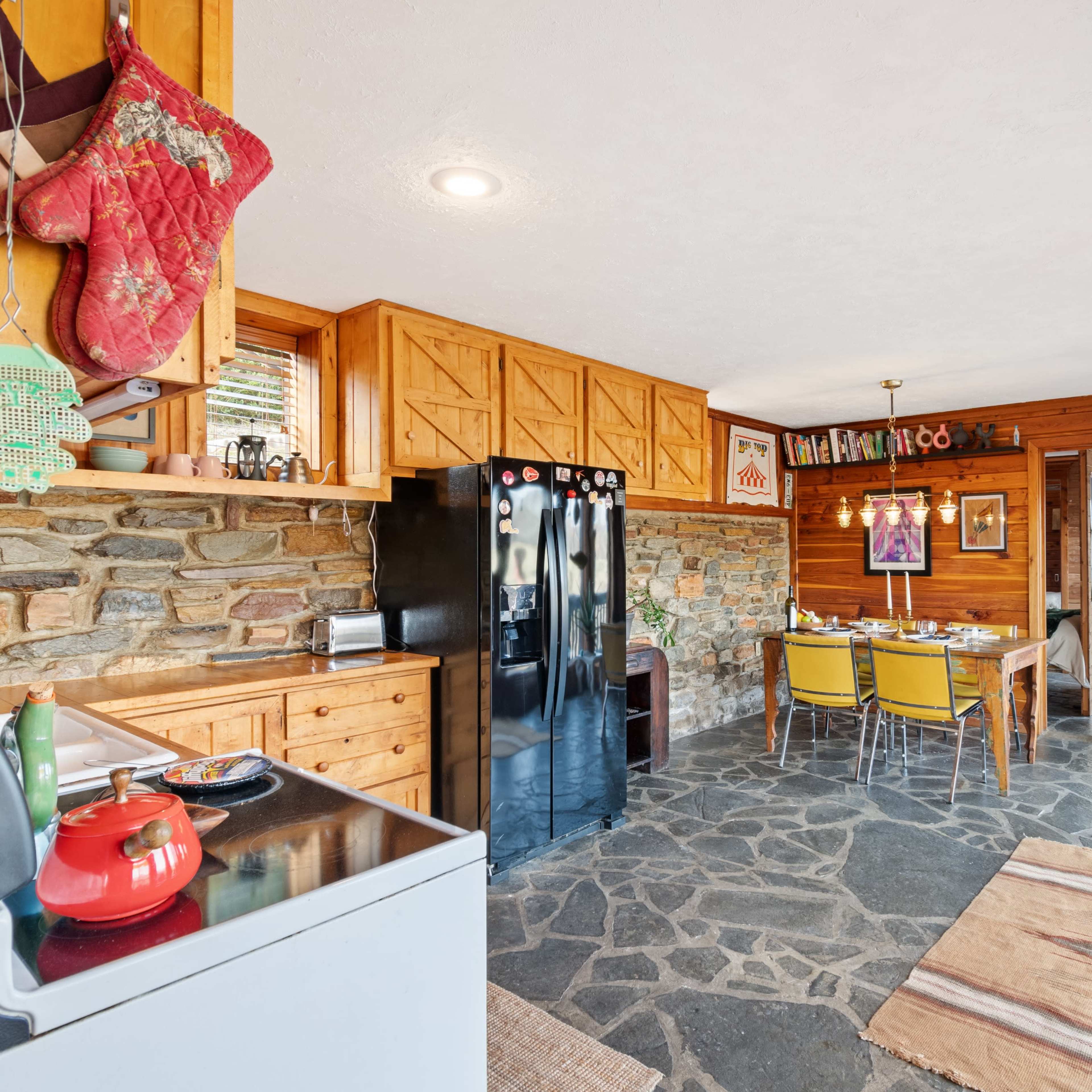 A kitchen and dining area featuring wooden cabinets, black refrigerators, a stone wall, and a table set with colorful chairs.