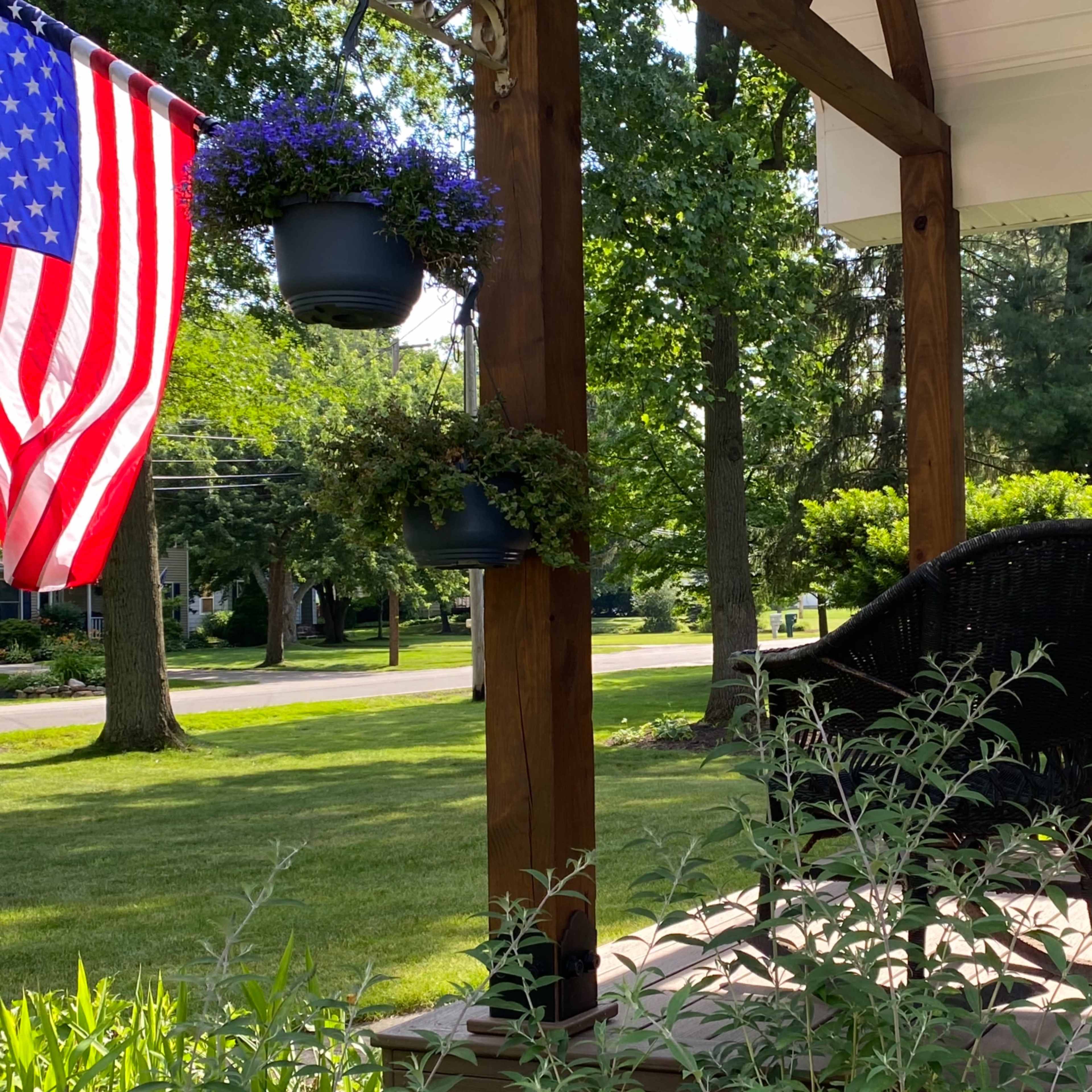A wicker chair is positioned on a wooden porch decorated with hanging flower pots, alongside an American flag.
