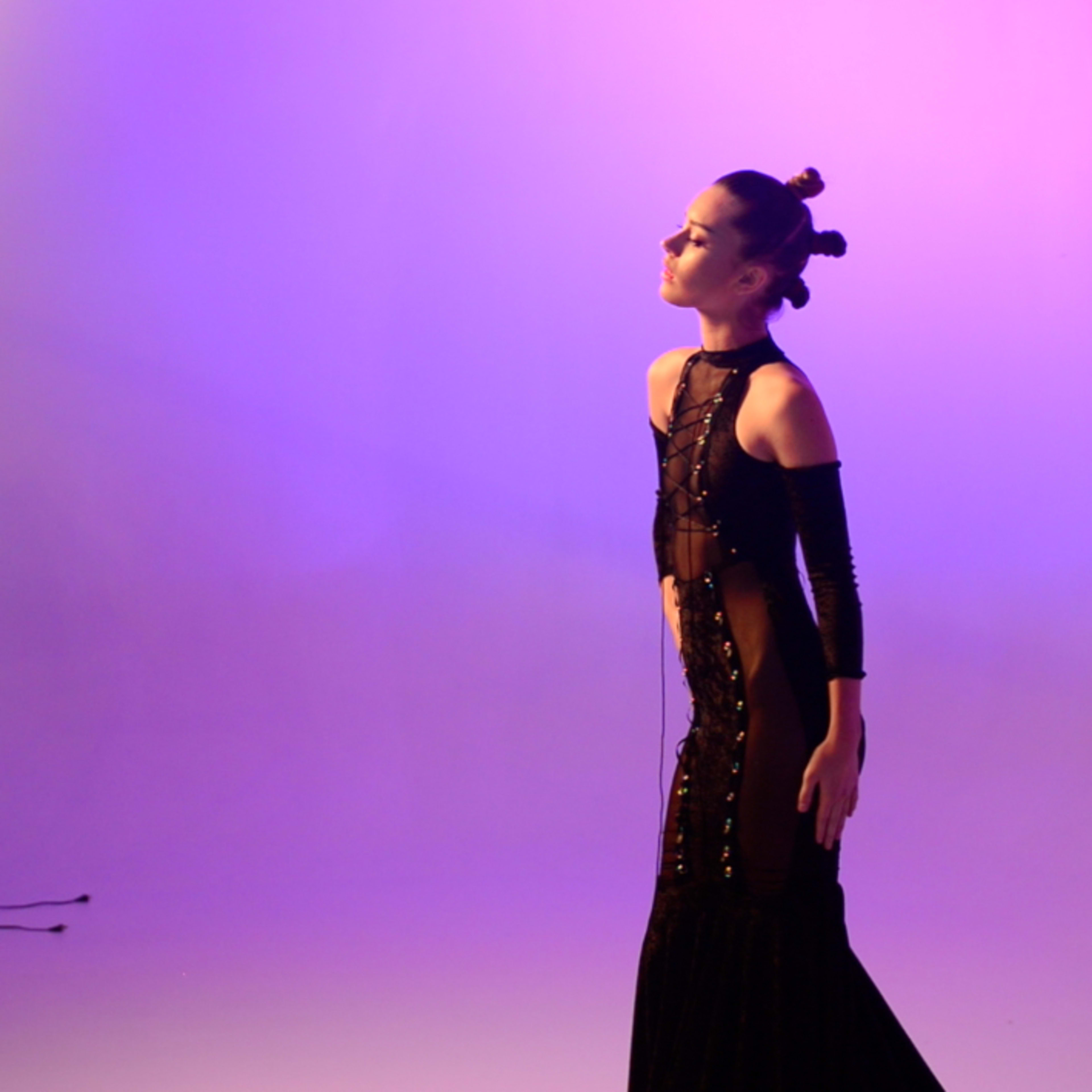 A model poses under a large studio light while wearing a black evening gown against a purple background.