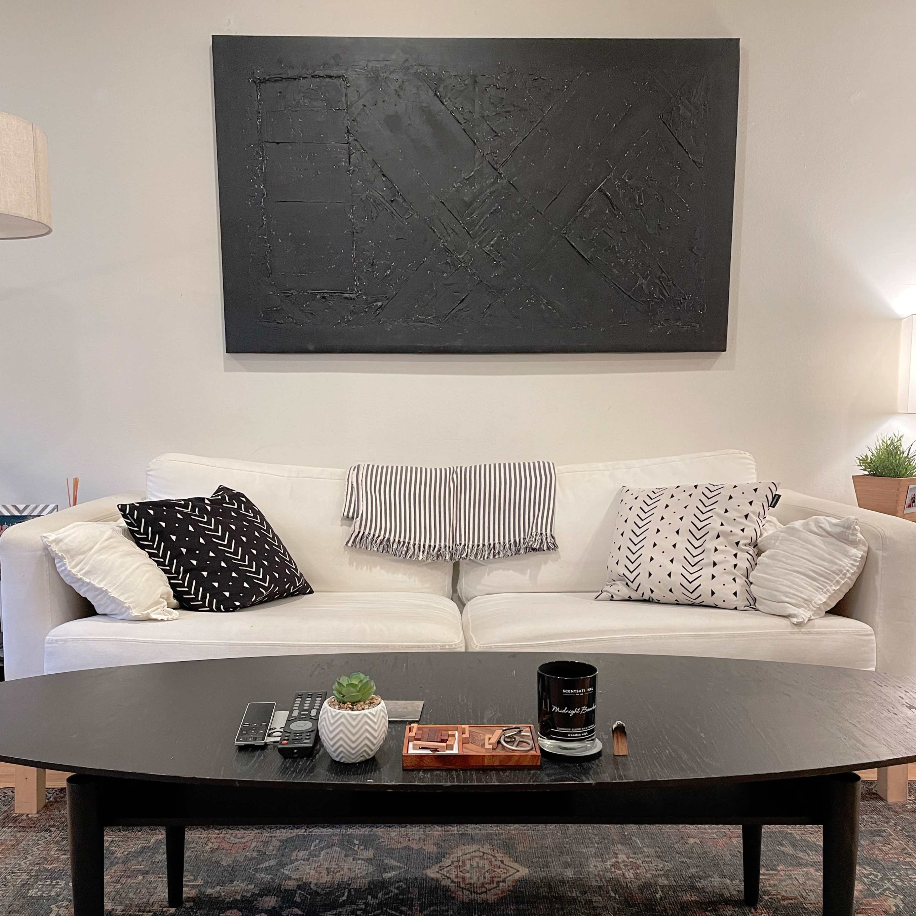 A white sofa with patterned cushions is positioned in front of a black coffee table on a geometric rug, with abstract wall art hanging above.