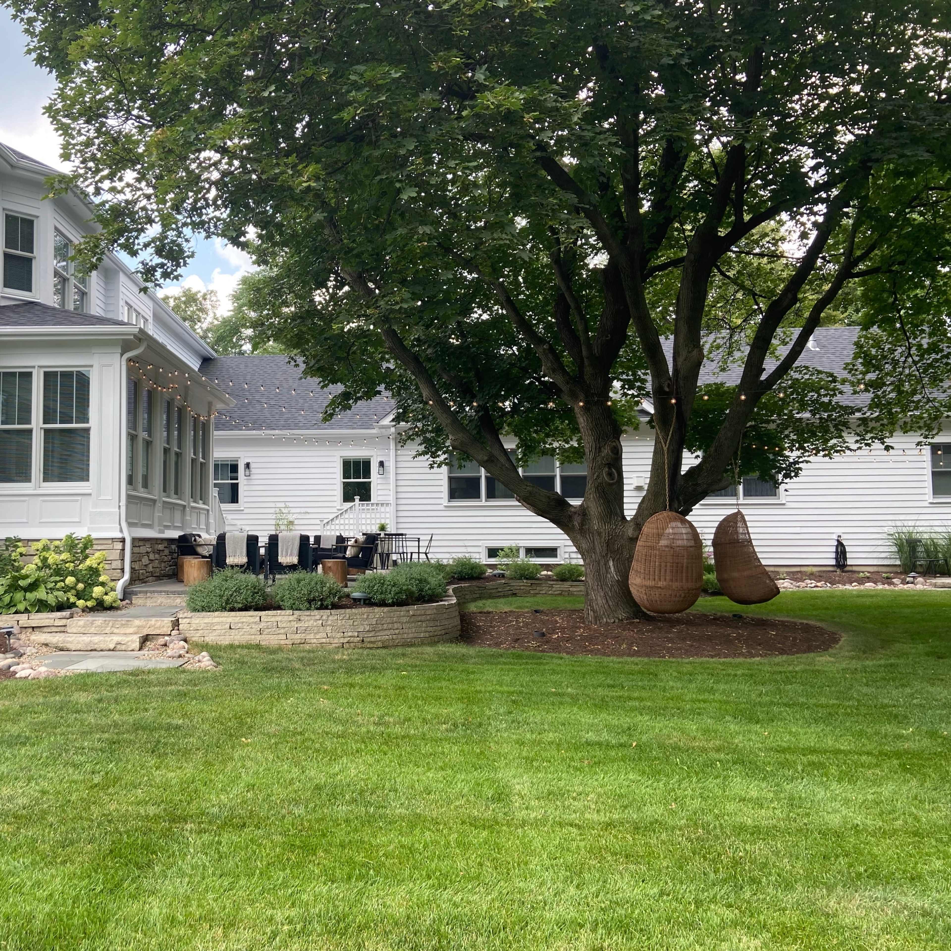 A white house with a large patio, surrounded by a landscaped yard featuring a tree and two hanging chairs.