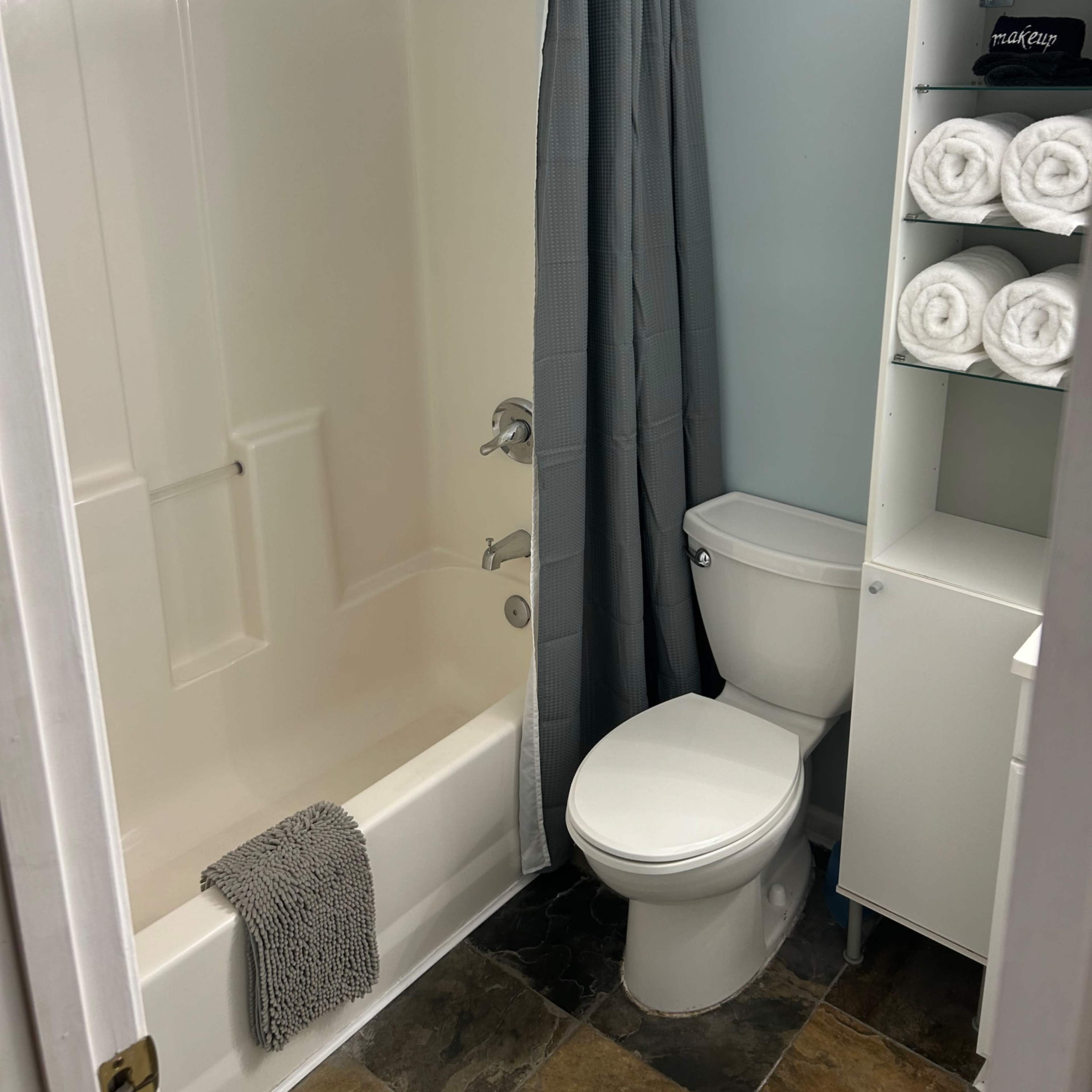 A bathroom with a bathtub-shower combination, a toilet, and a storage unit with neatly stacked towels.