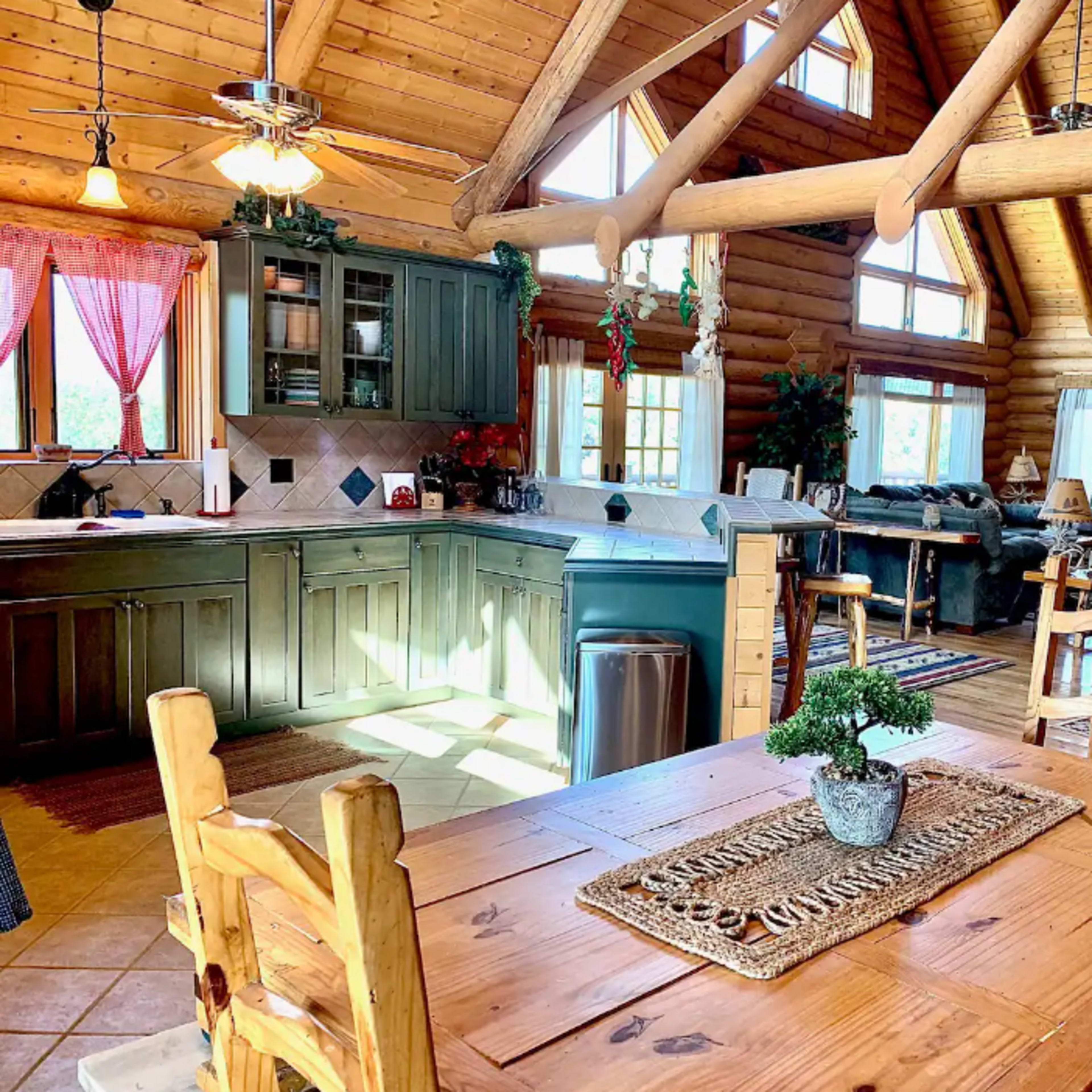 A wooden kitchen and dining area in a log cabin, featuring a table, chairs, and large windows allowing natural light.