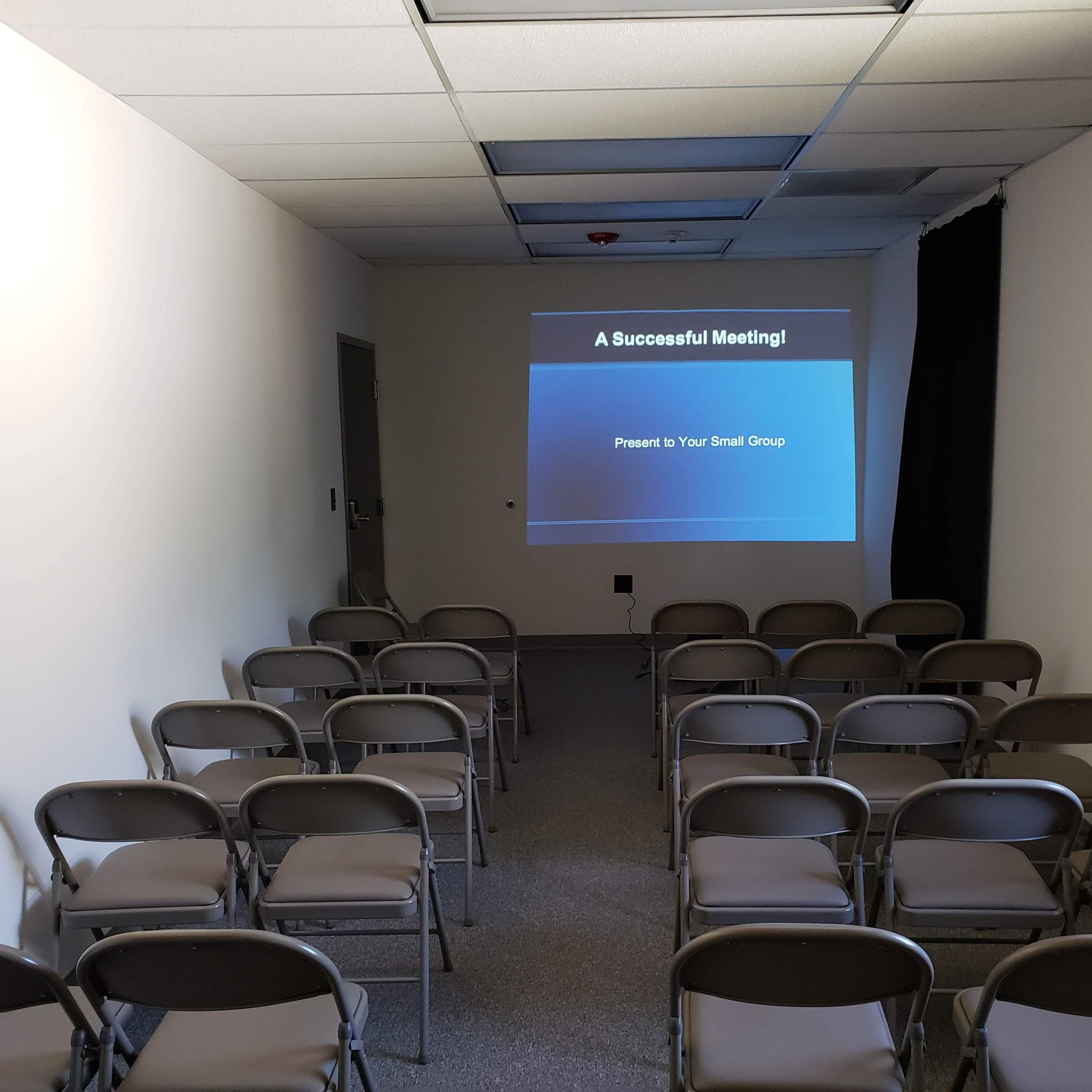 A small, empty room arranged with rows of folding chairs facing a table, with a window and a television in the corner.