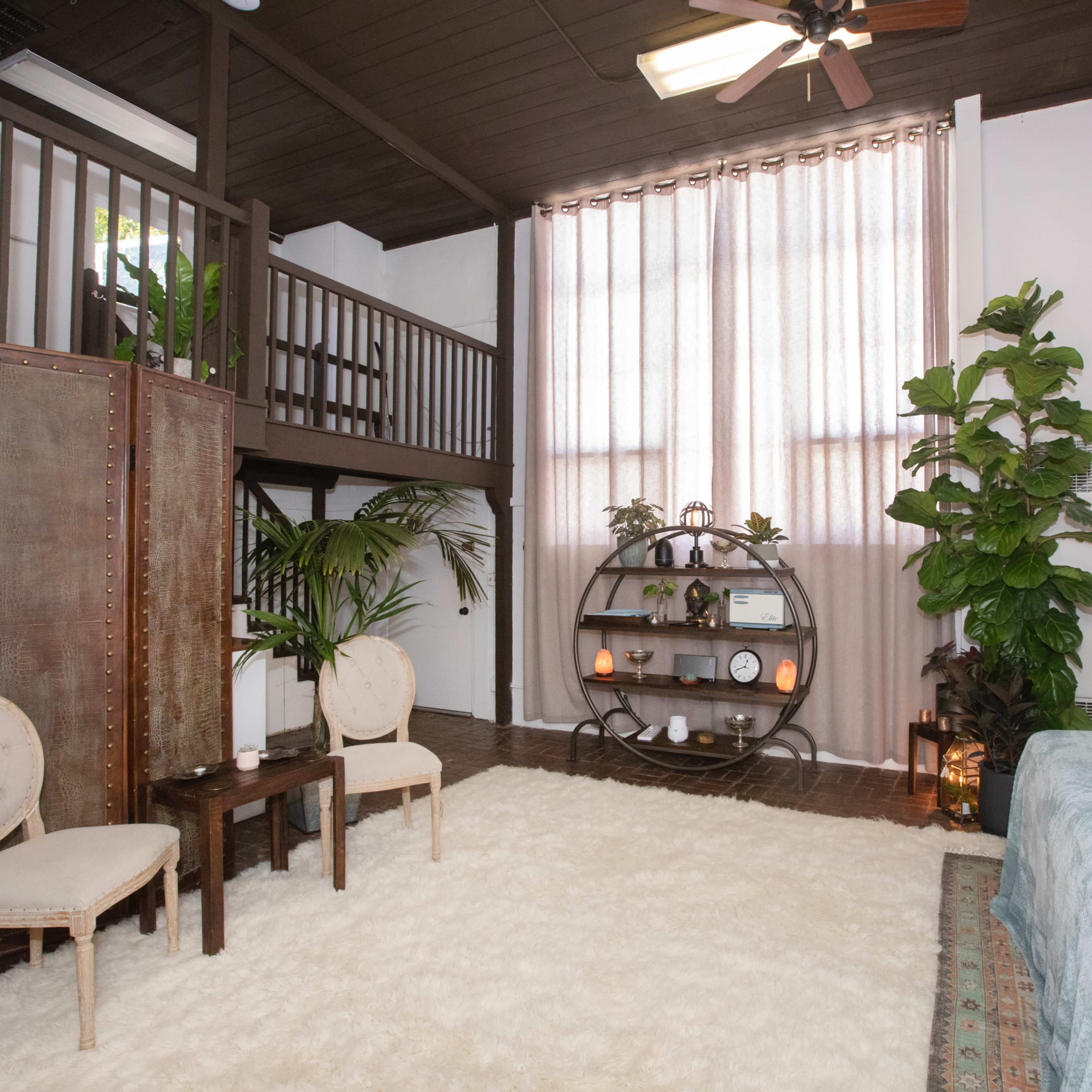 A well-lit room featuring a cozy seating area with two chairs and a small table, a large bed, decorative plants, and a tall window with sheer curtains.