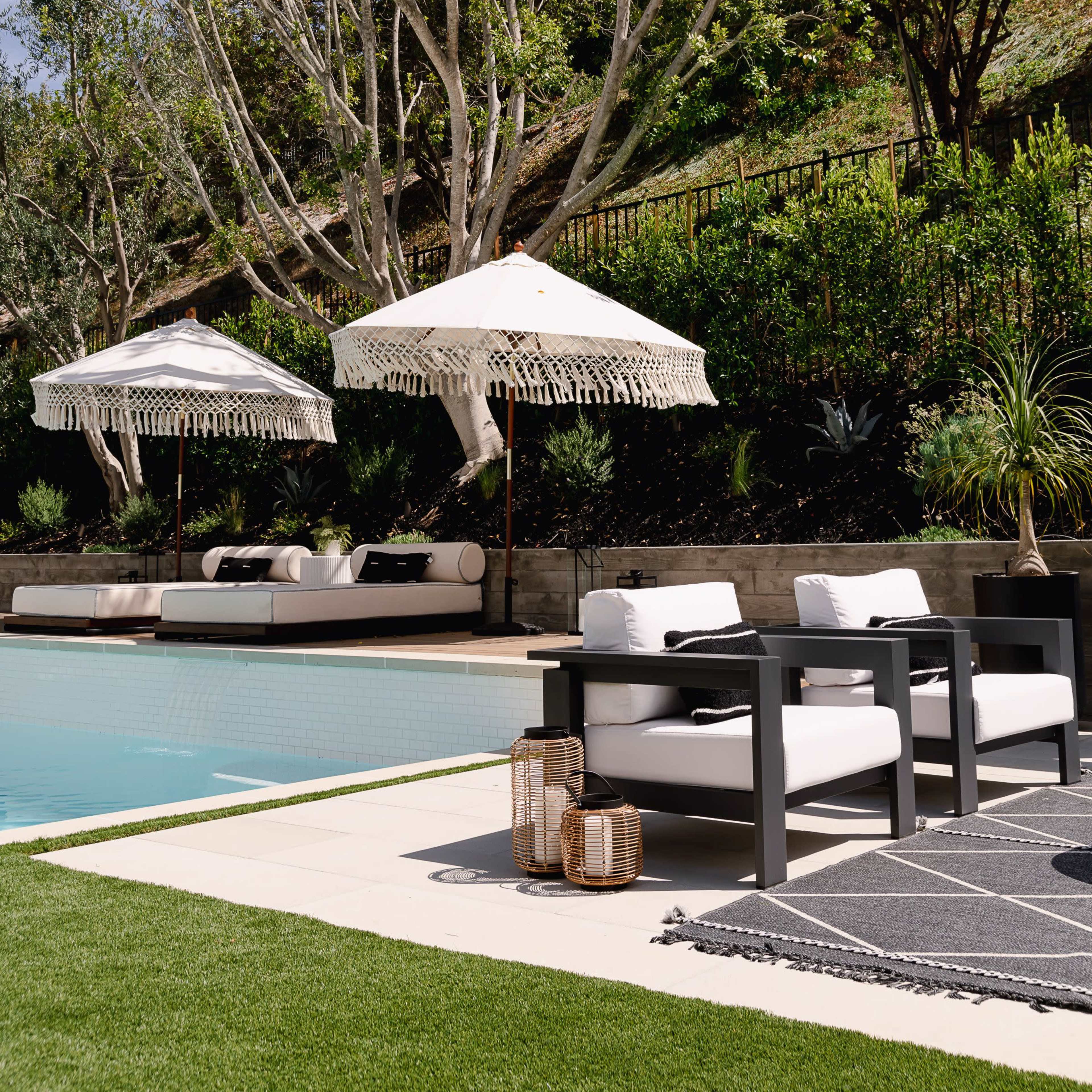 A modern outdoor pool area with two white lounge chairs, patterned rugs, and large umbrellas beside a well-maintained garden.