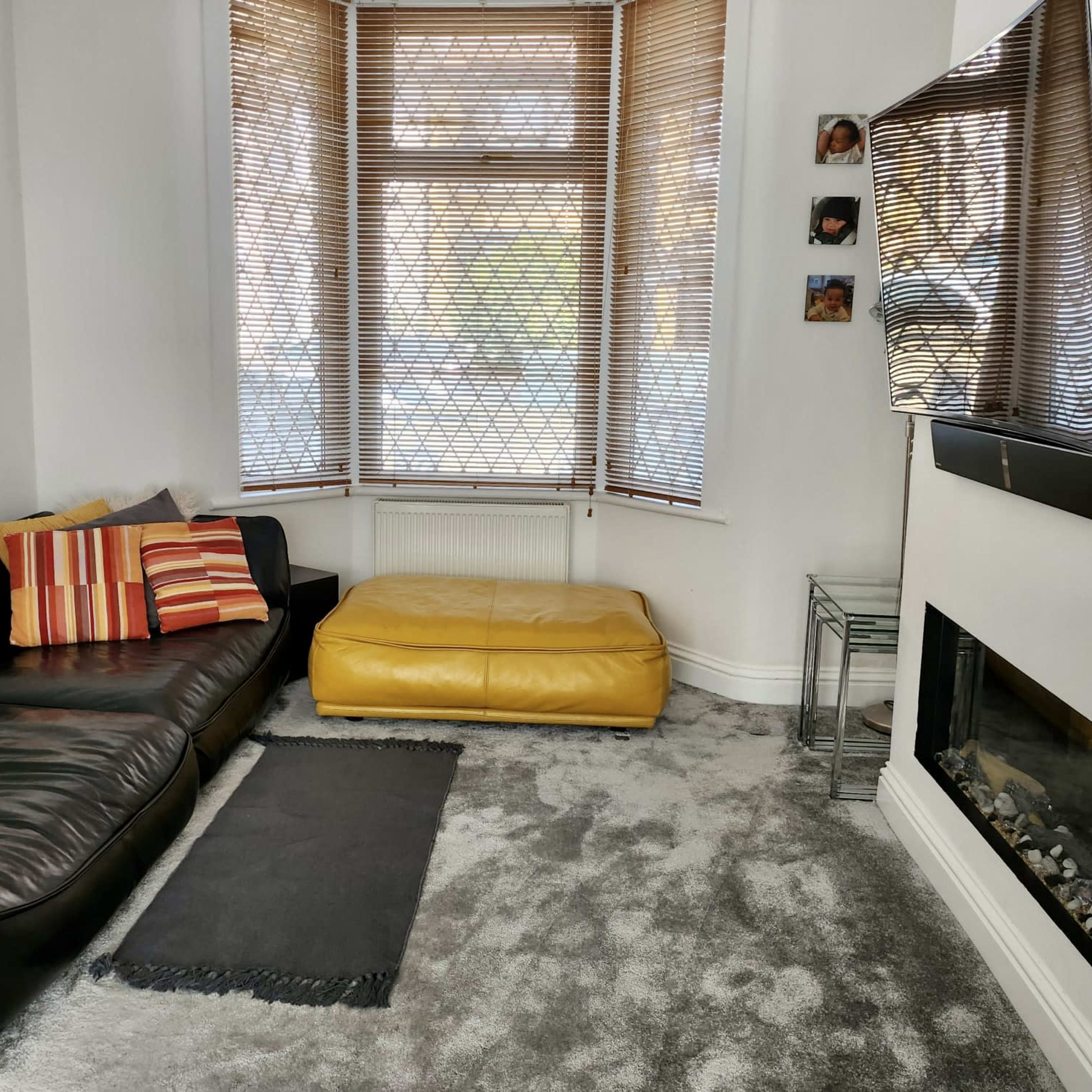 A living room featuring a black leather sofa with striped cushions, a yellow ottoman, a television mounted on the wall, and a fireplace below it, with large windows covered by wooden blinds.