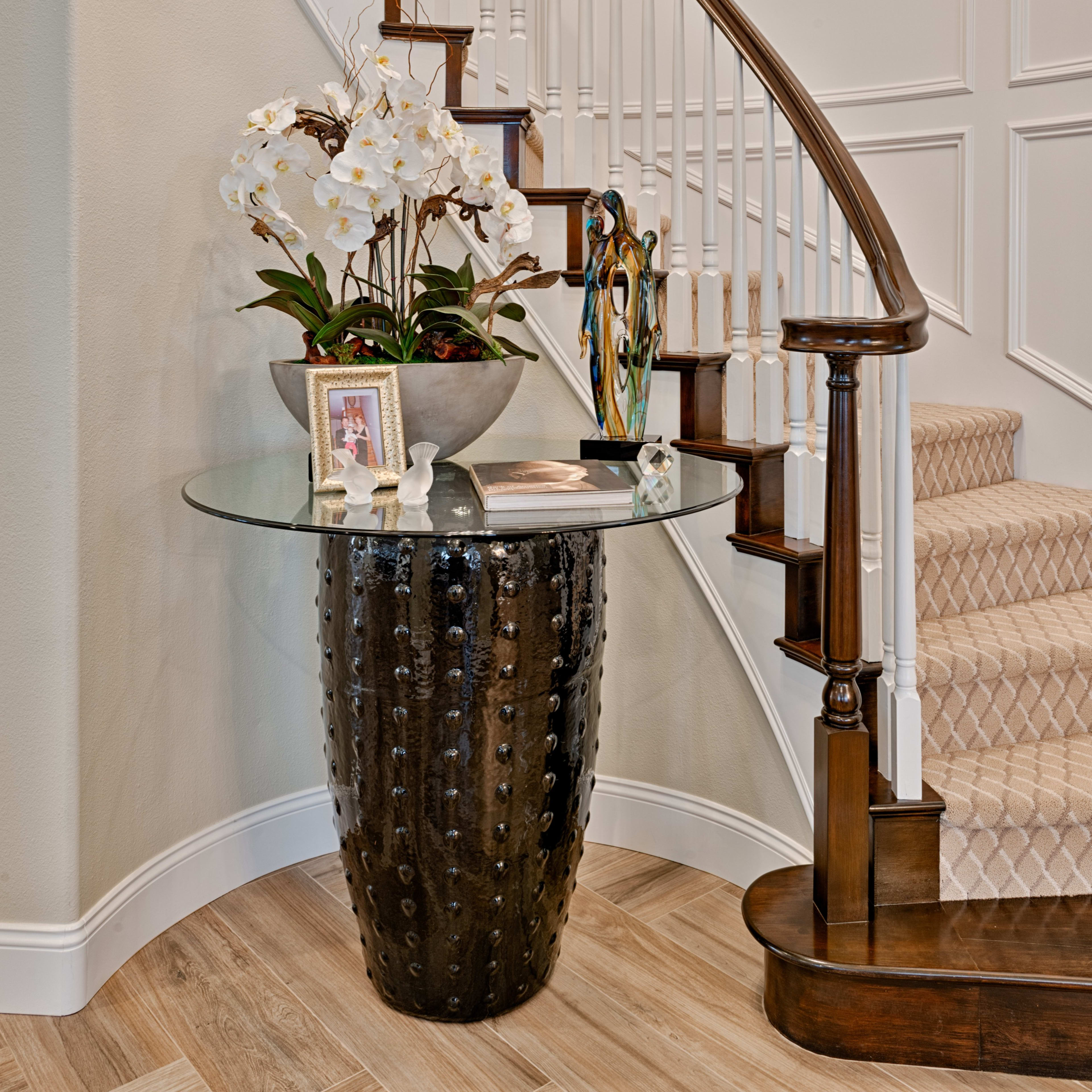 A curved staircase with wooden railings surrounds a decorative table featuring a glass top and a floral arrangement, positioned in a well-lit foyer.