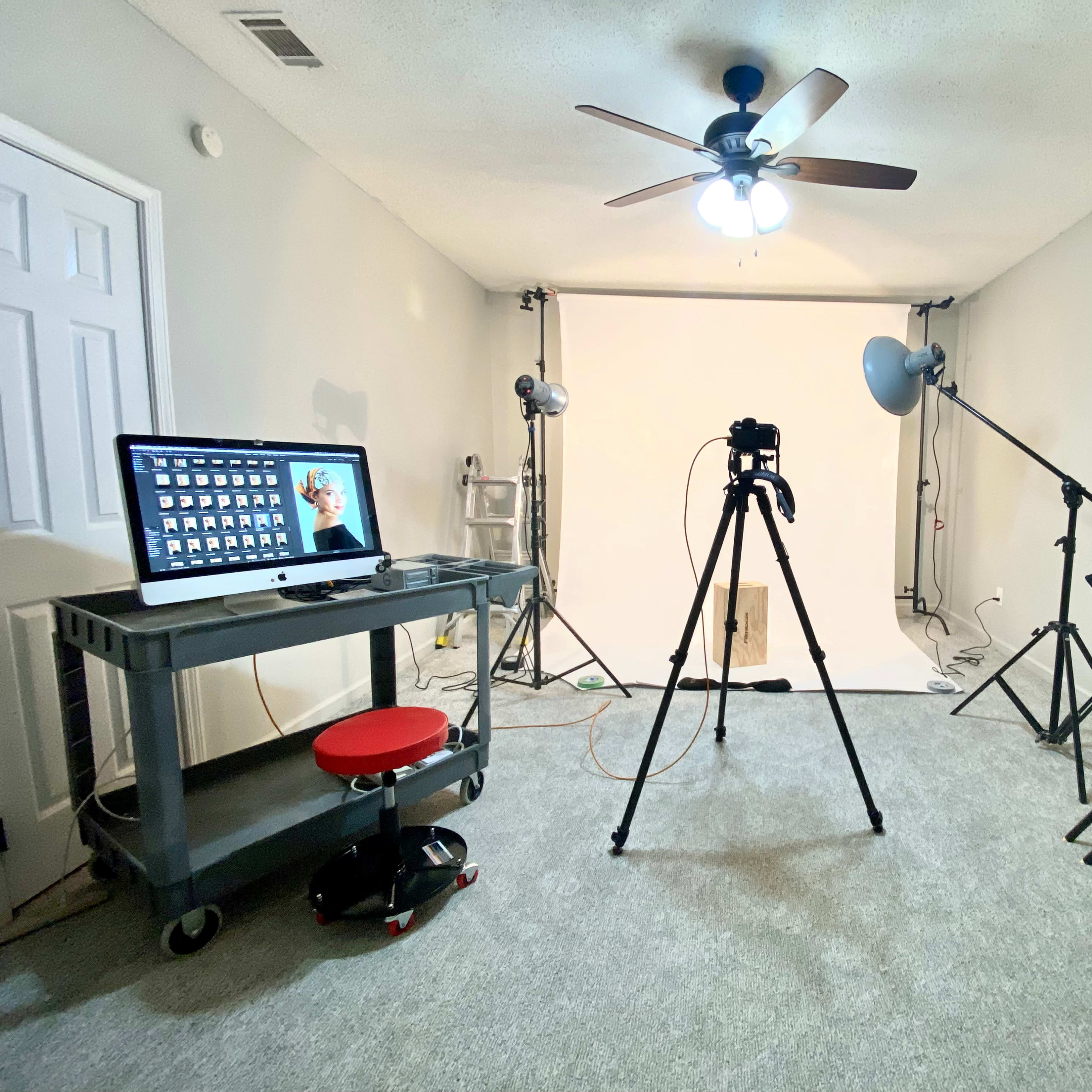A photography studio is set up with a computer, a camera on a tripod, and several lighting equipment stands against a plain backdrop.