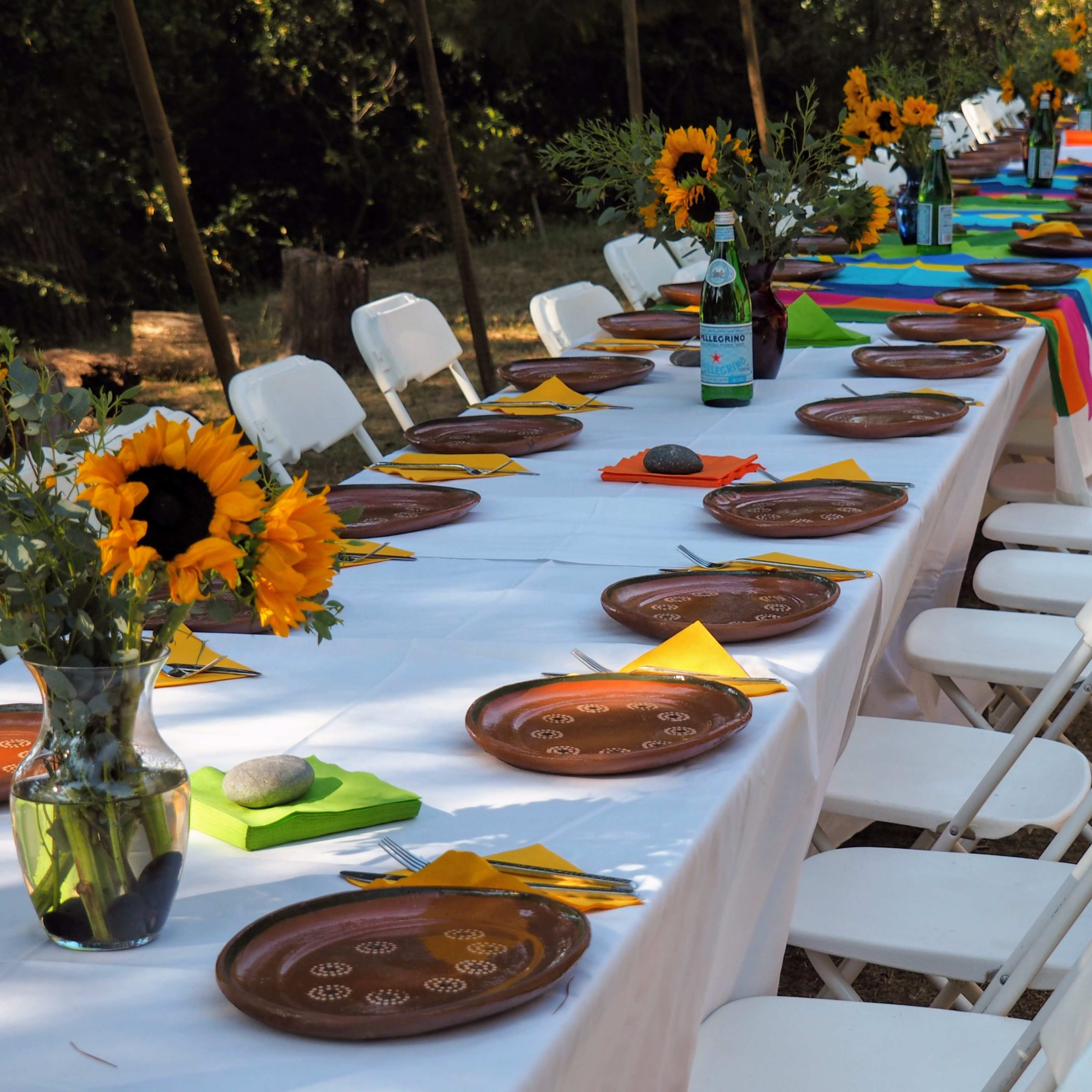 A long outdoor table is set with brown plates, colorful napkins, and vases of sunflowers, arranged beneath a canopy.