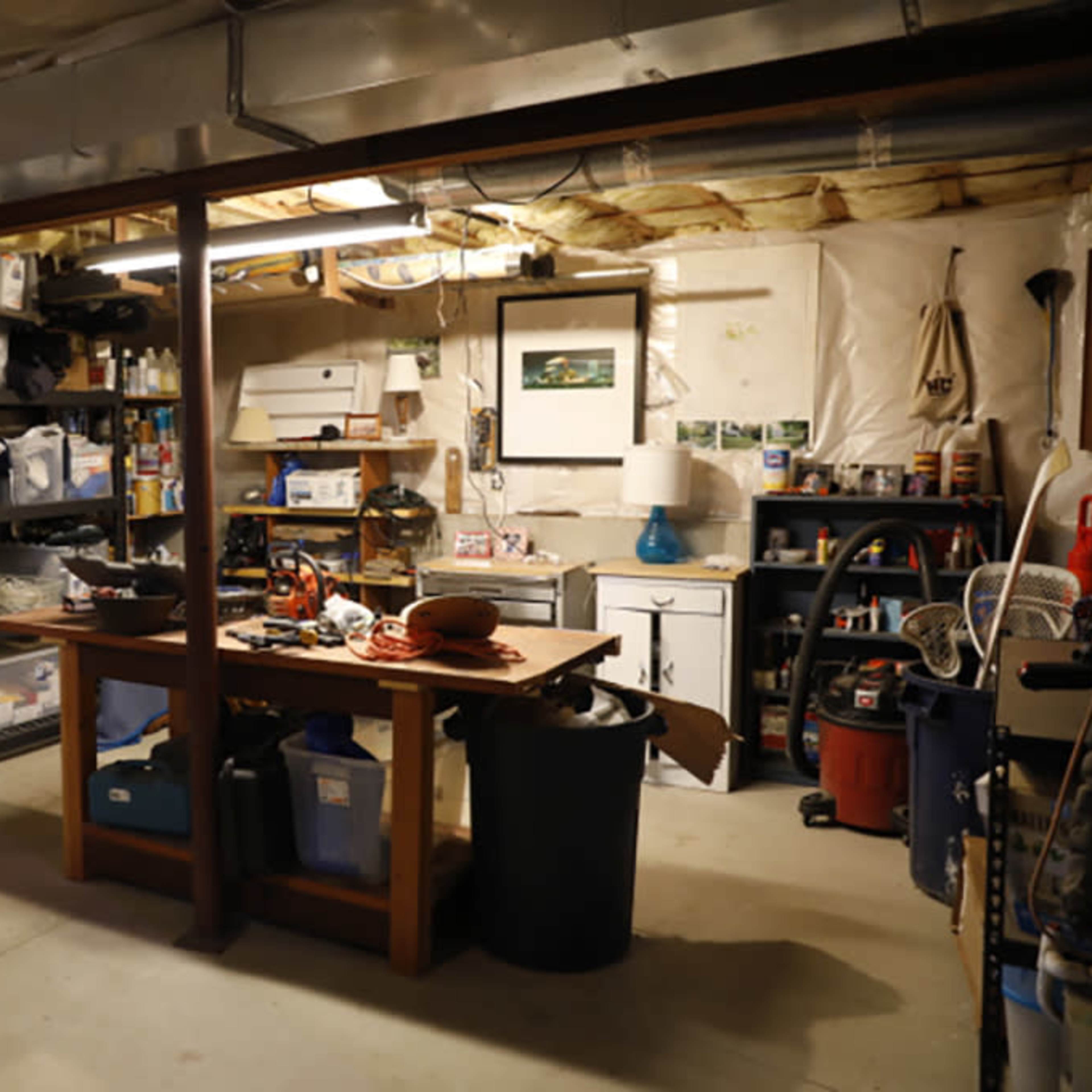 A basement workspace with shelves filled with boxes and tools, a central table, and a lamp.