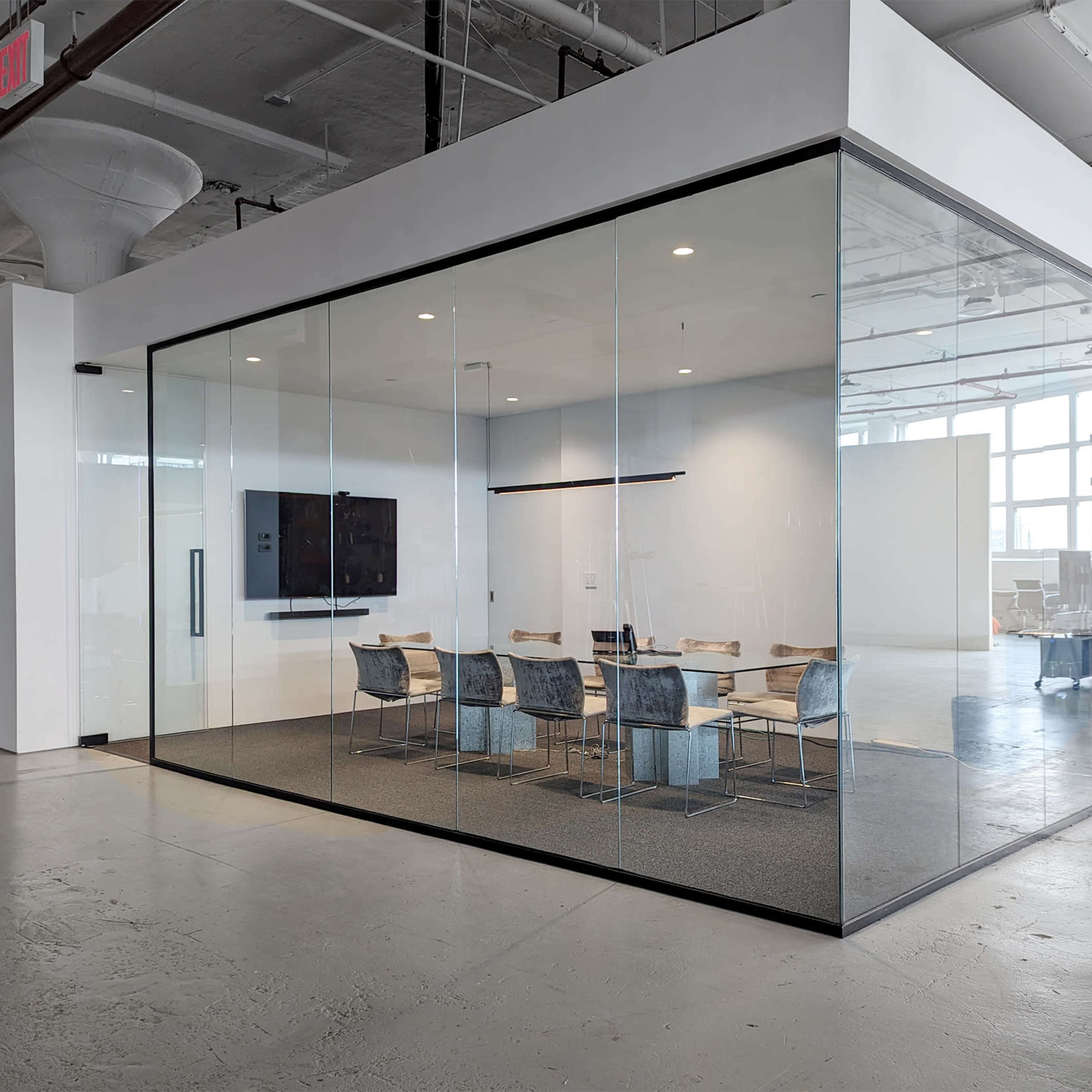 A modern conference room enclosed in clear glass walls, featuring a large table with chairs and a wall-mounted screen.